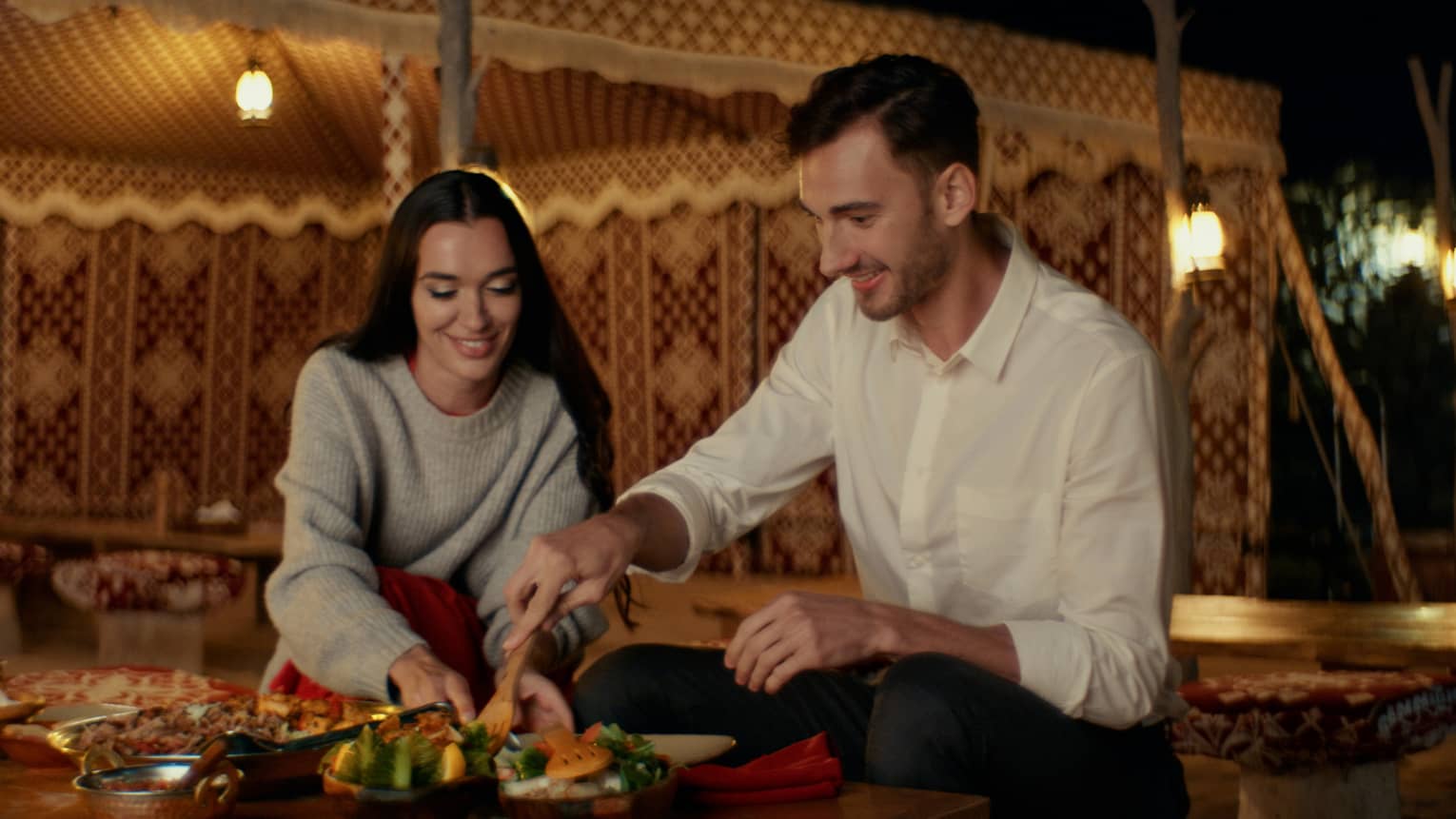 A couple in front of a buffet of food in a lavish tent, grabbing a green dish.