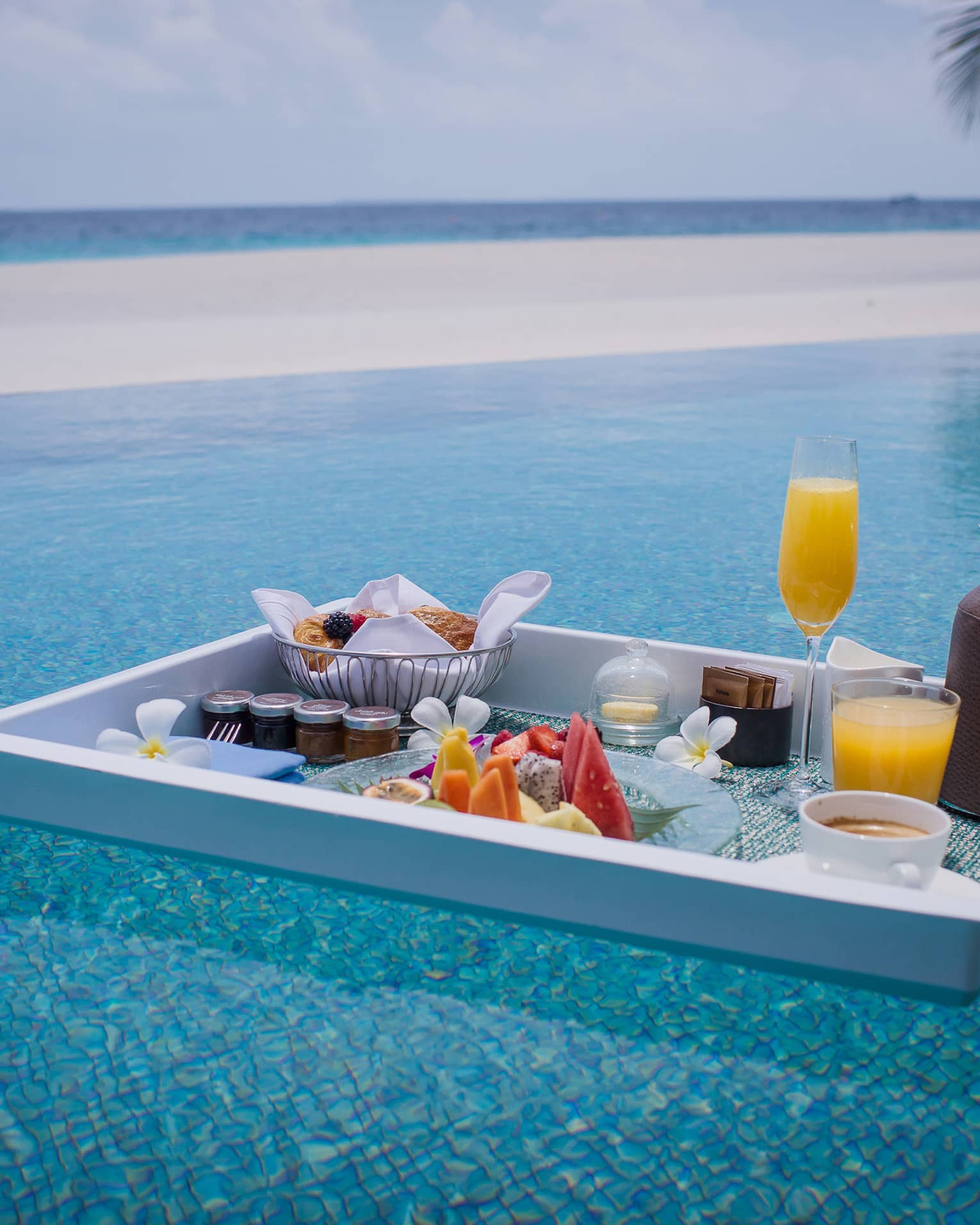 Floating in a shimmering turquoise pool, is a white tray with a selection of fruit, pastries, jams, beverages and flowers.