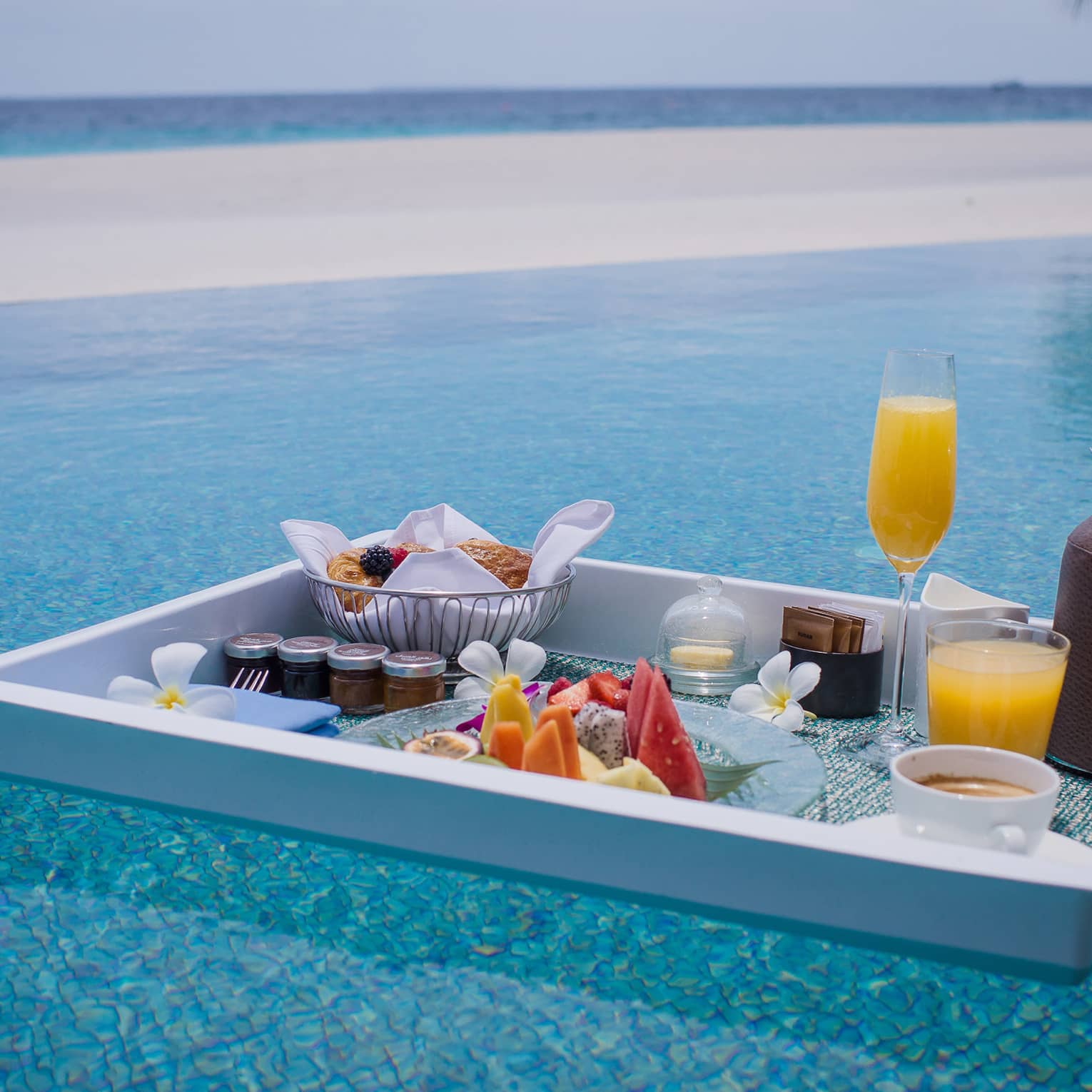 Floating in a shimmering turquoise pool, is a white tray with a selection of fruit, pastries, jams, beverages and flowers.