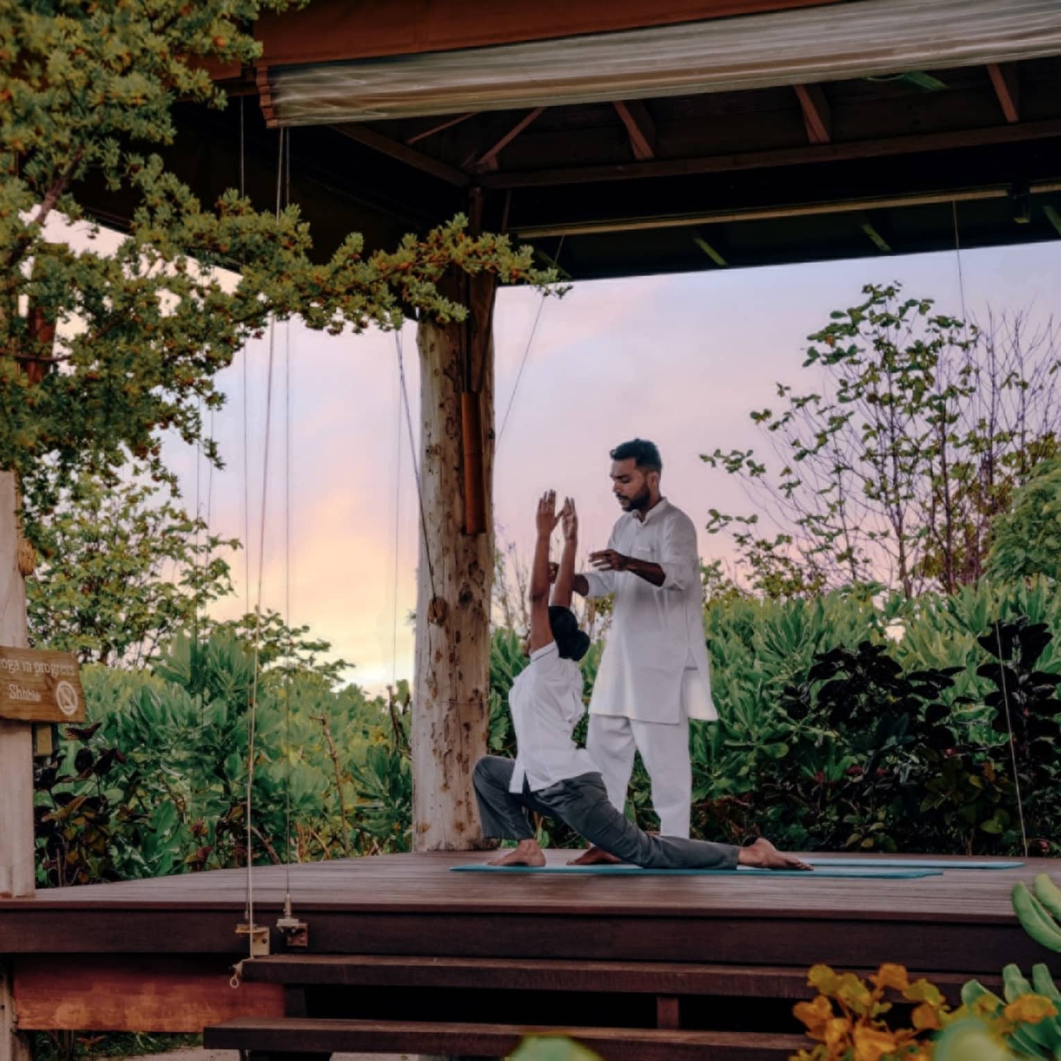Instructor guides student doing yoga in an open-air pavilion surrounded by greenery
