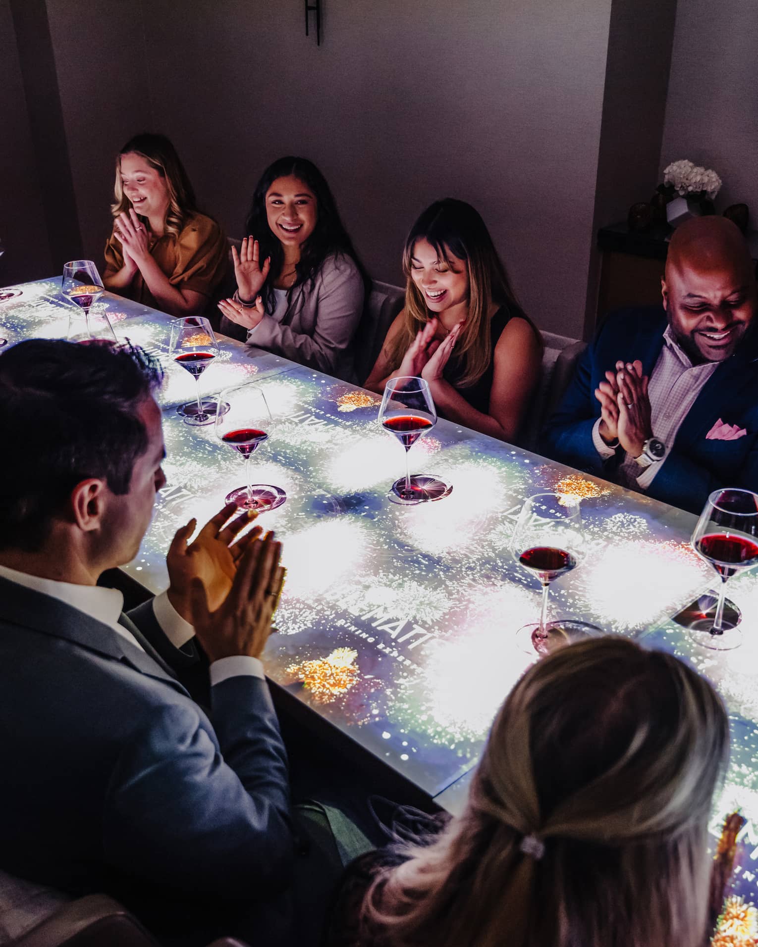 A large group of people sitting at a table with a screen on top of it drinking wine.