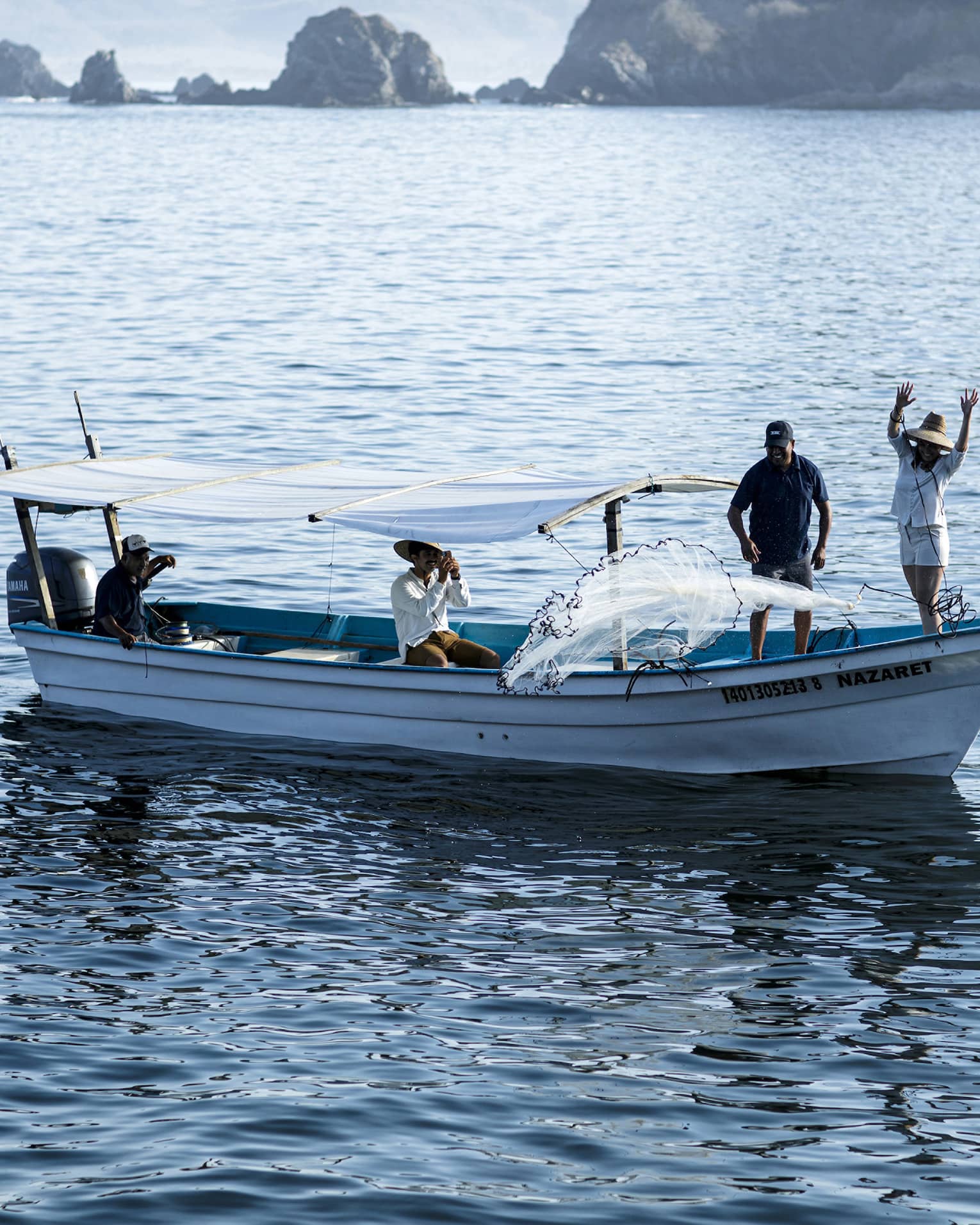 Side view of a boat with two people at the bow casting a net into the water, two others seated under a canopy near the stern.