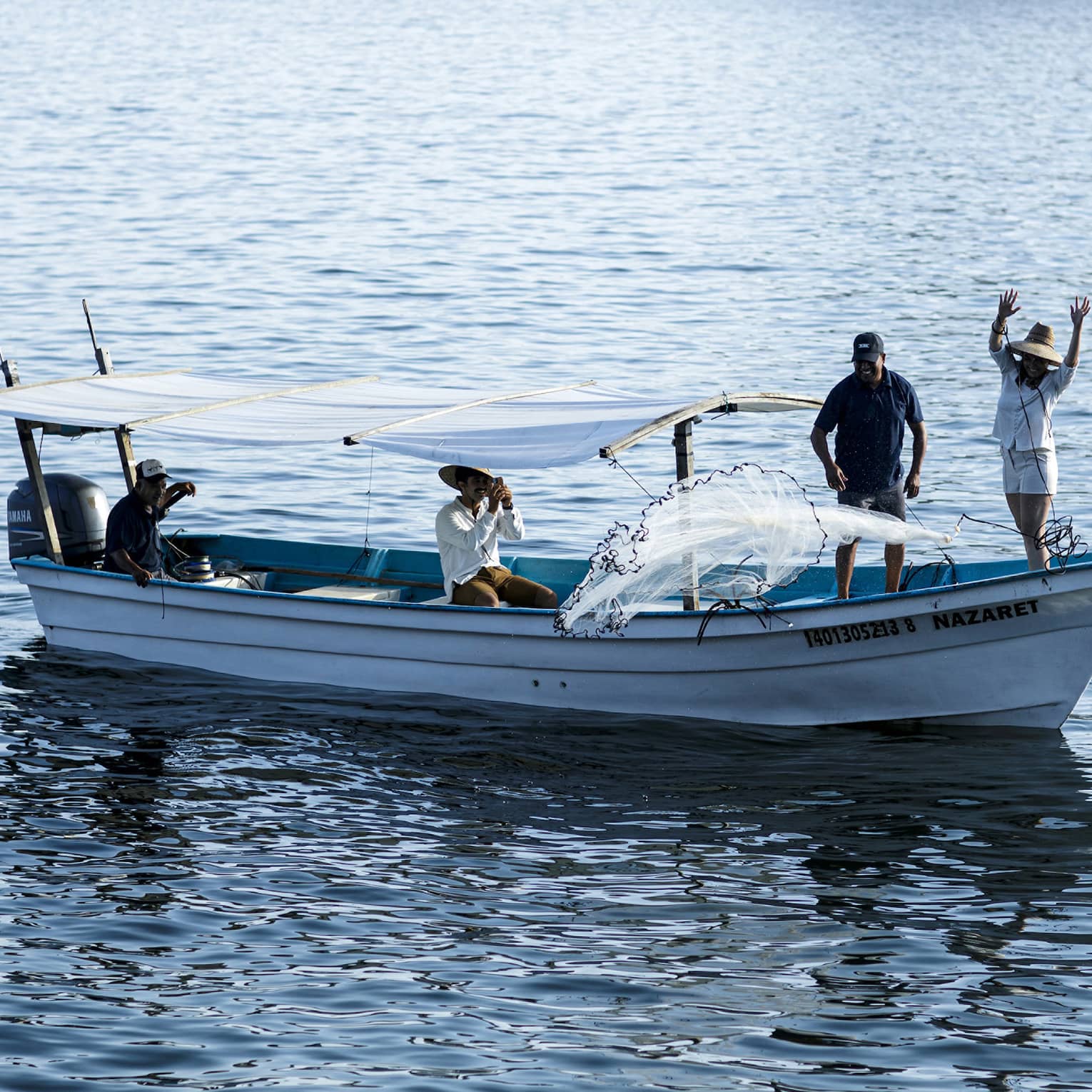 Side view of a boat with two people at the bow casting a net into the water, two others seated under a canopy near the stern.