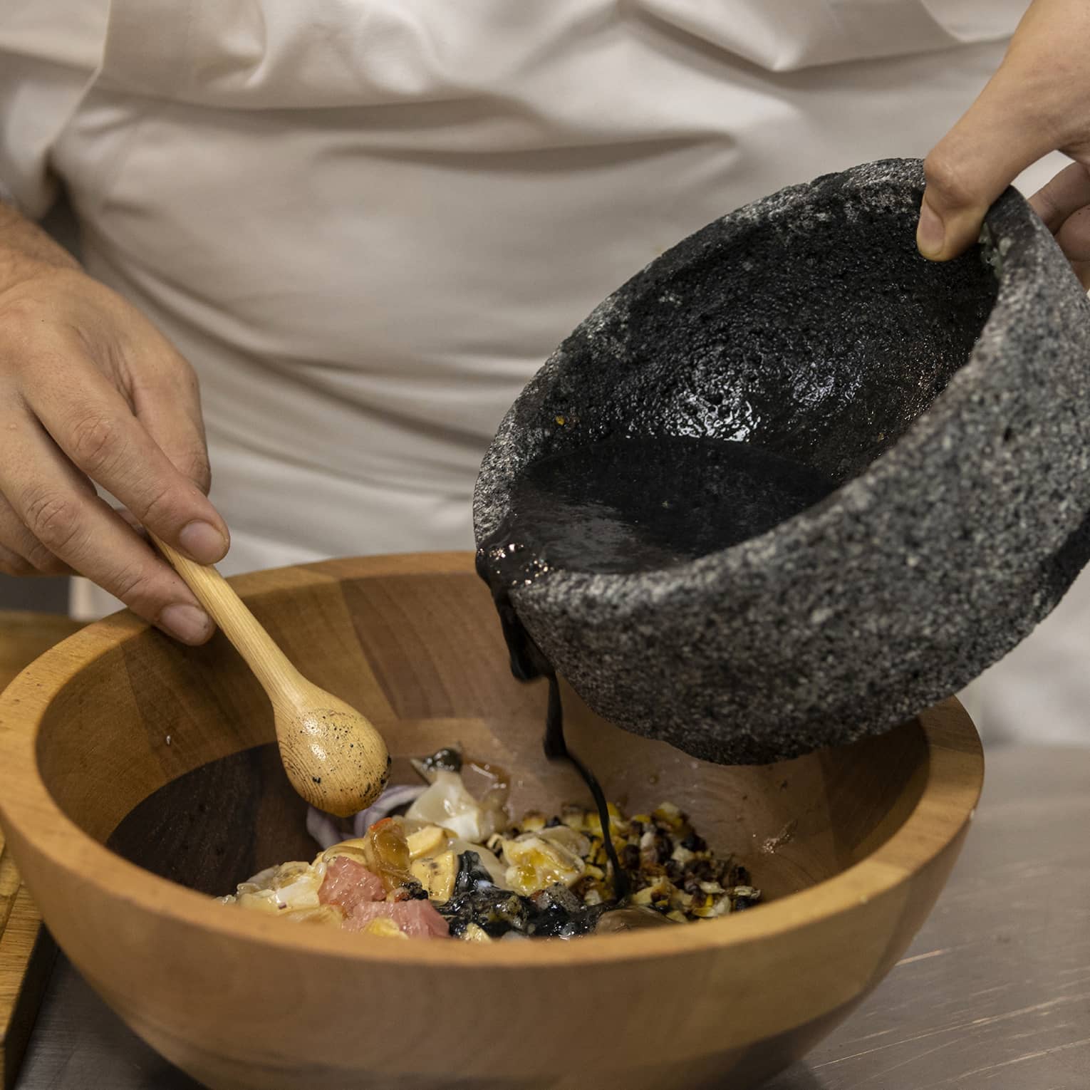 Chef pours black sauce from a stone bowl into a wood bowl of ingredients, a small wooden spoon in the other hand. 
