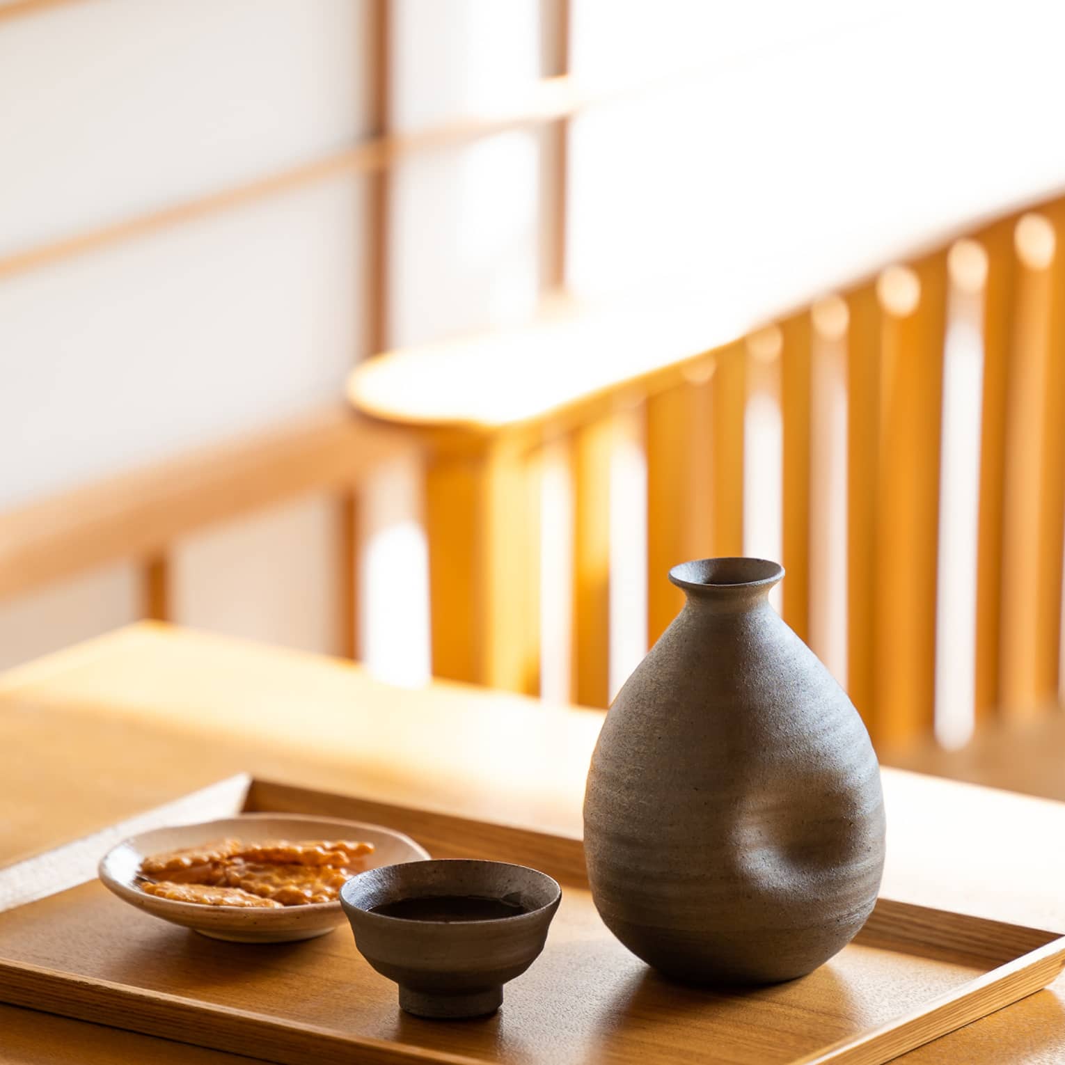Close-up of a dark ceramic sake carafe with matching cup and a small plate of crackers on a wooden tray and table.