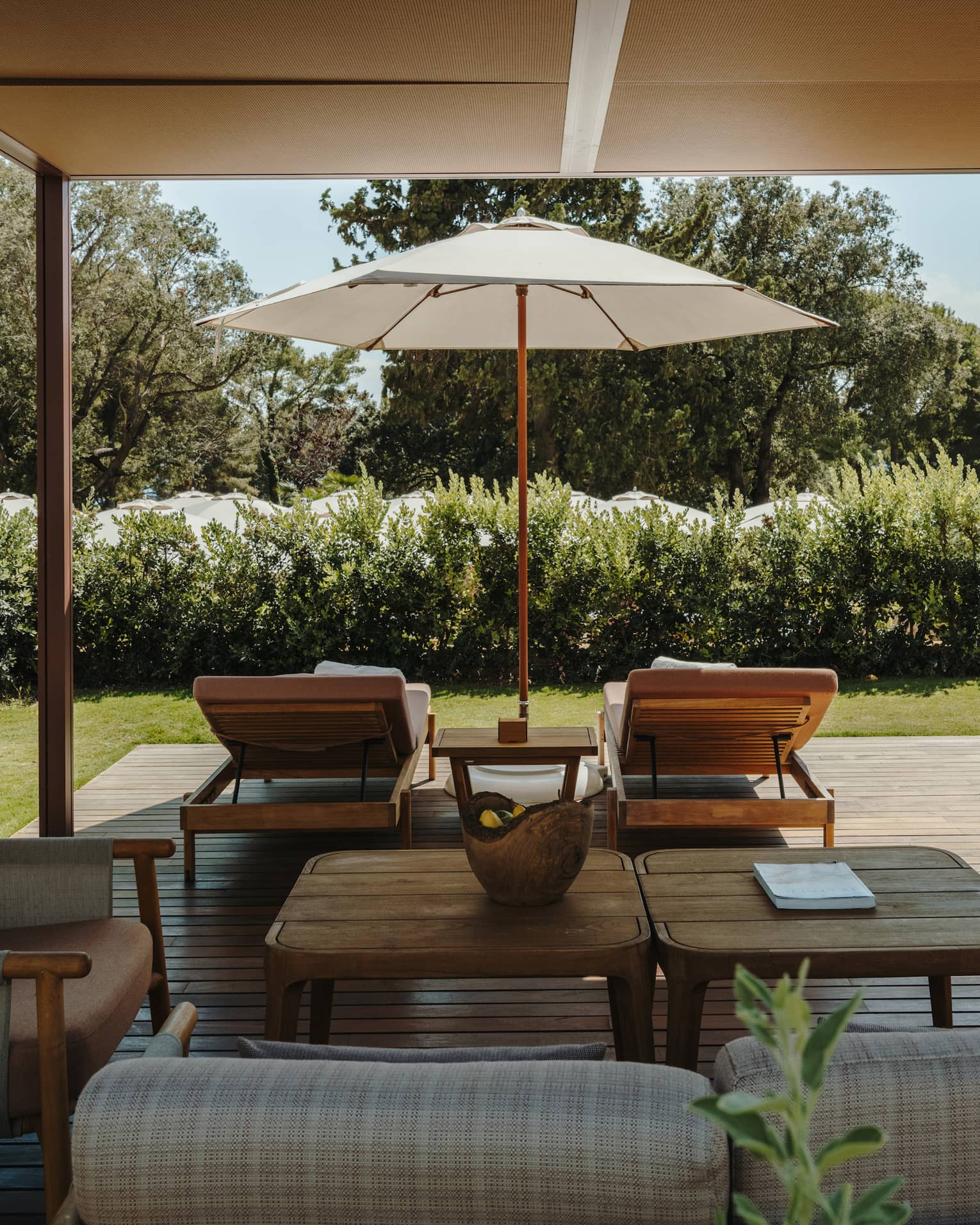 A shaded pool cabana with comfortable seating, a table, lounge chairs, an umbrella and lush green surroundings.