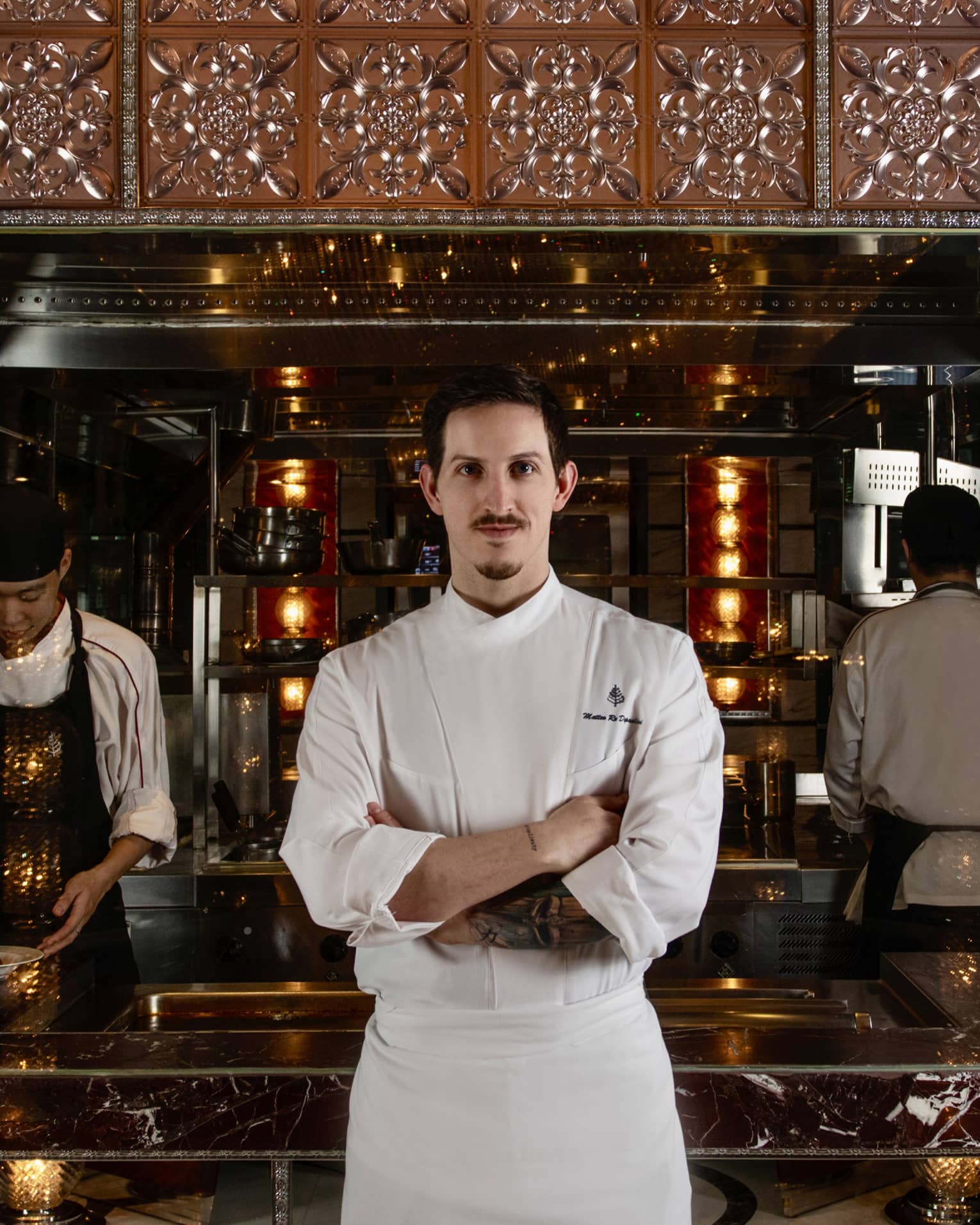 Chef stands, arms crossed, facing the camera in front of a marble counter where two chefs prepare food