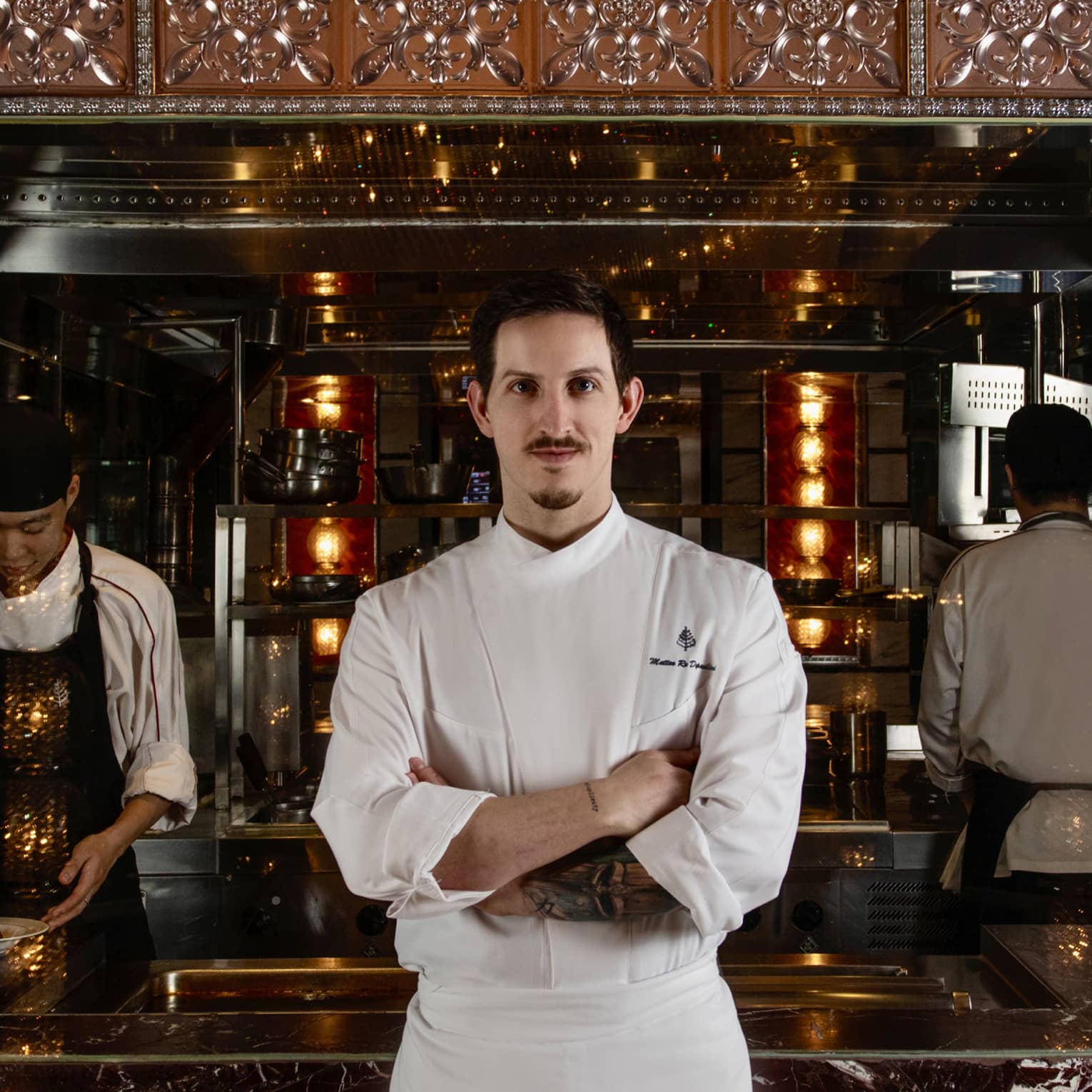 Chef stands, arms crossed, facing the camera in front of a marble counter where two chefs prepare food