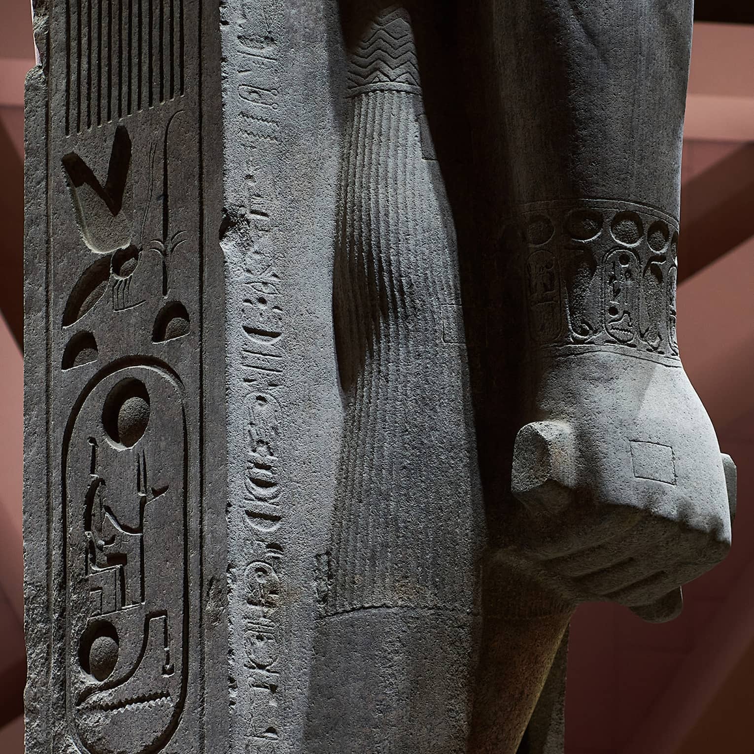 Close-up view of a statue's hand in a fist, supported by a stone inscribed with hyroglyph carvings. 