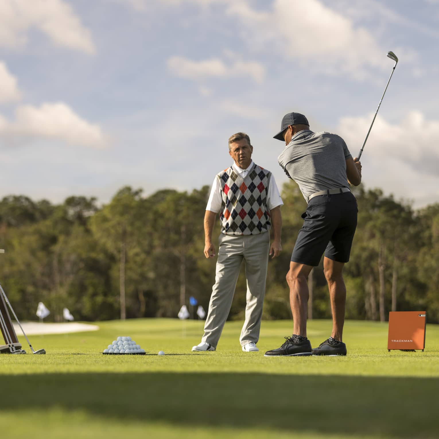 Against clear sky, one golfer looks on as another prepares to swing, a tray of balls and a stand of clubs nearby.