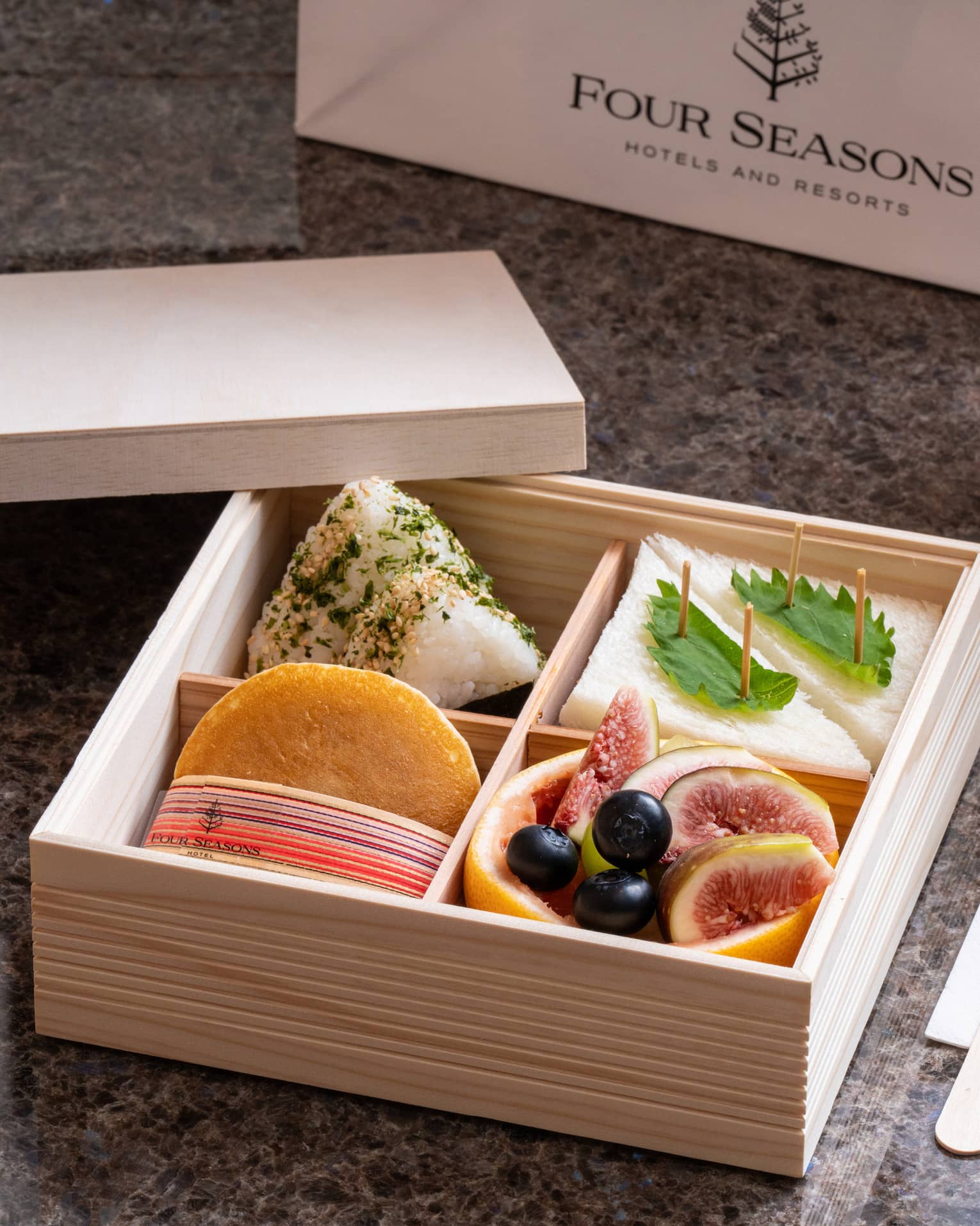 A wooden Four Seasons bento box with four compartments holding sandwiches, fruits and delicacies, beverage bottles nearby.