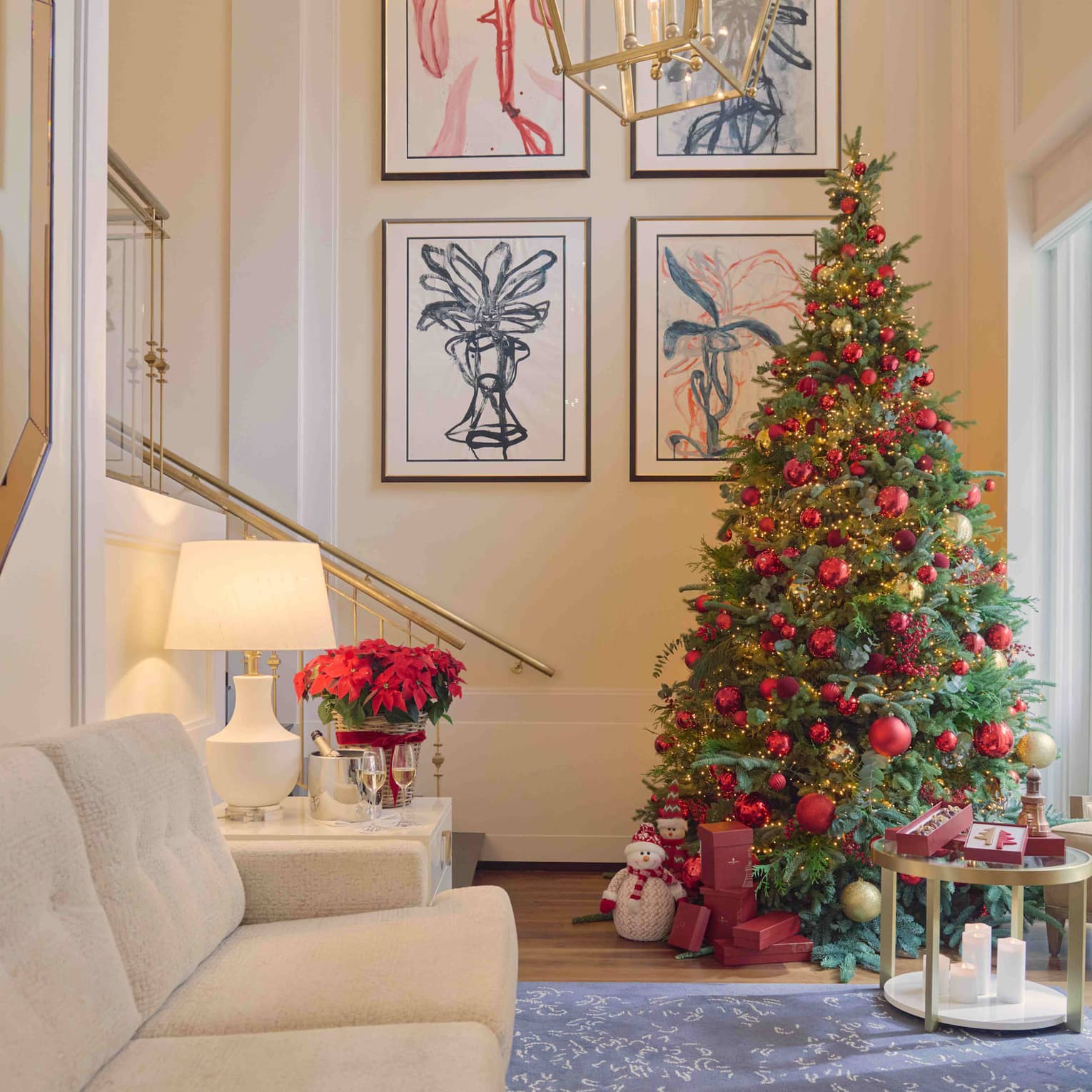 A spacious guest room with pale neutral decor, an off-white couch, an end table with a table lamp and vase of flowers, an upholstered chair, a large mirror and wall art. A decorated Christmas tree stands in a corner.
