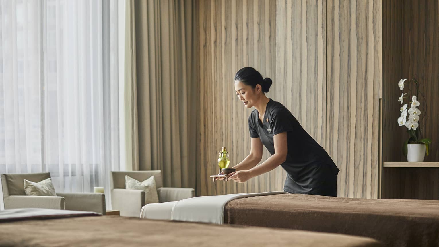 A female masseuse brings a drink to sit behind one of two massage beds. 