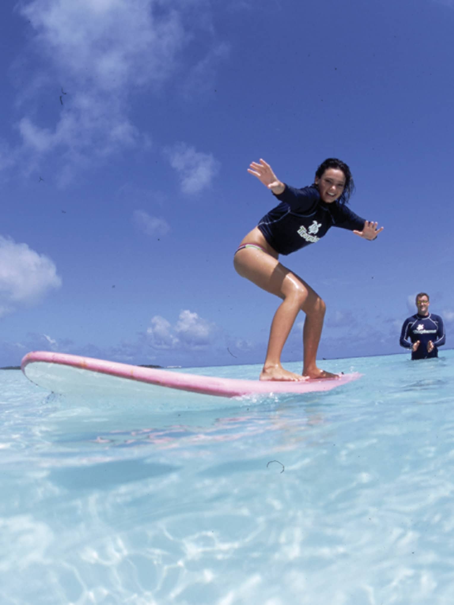 In the crystal-clear shimmering water is a woman learning to surf and an instructor standing in waist-deep water clapping.
