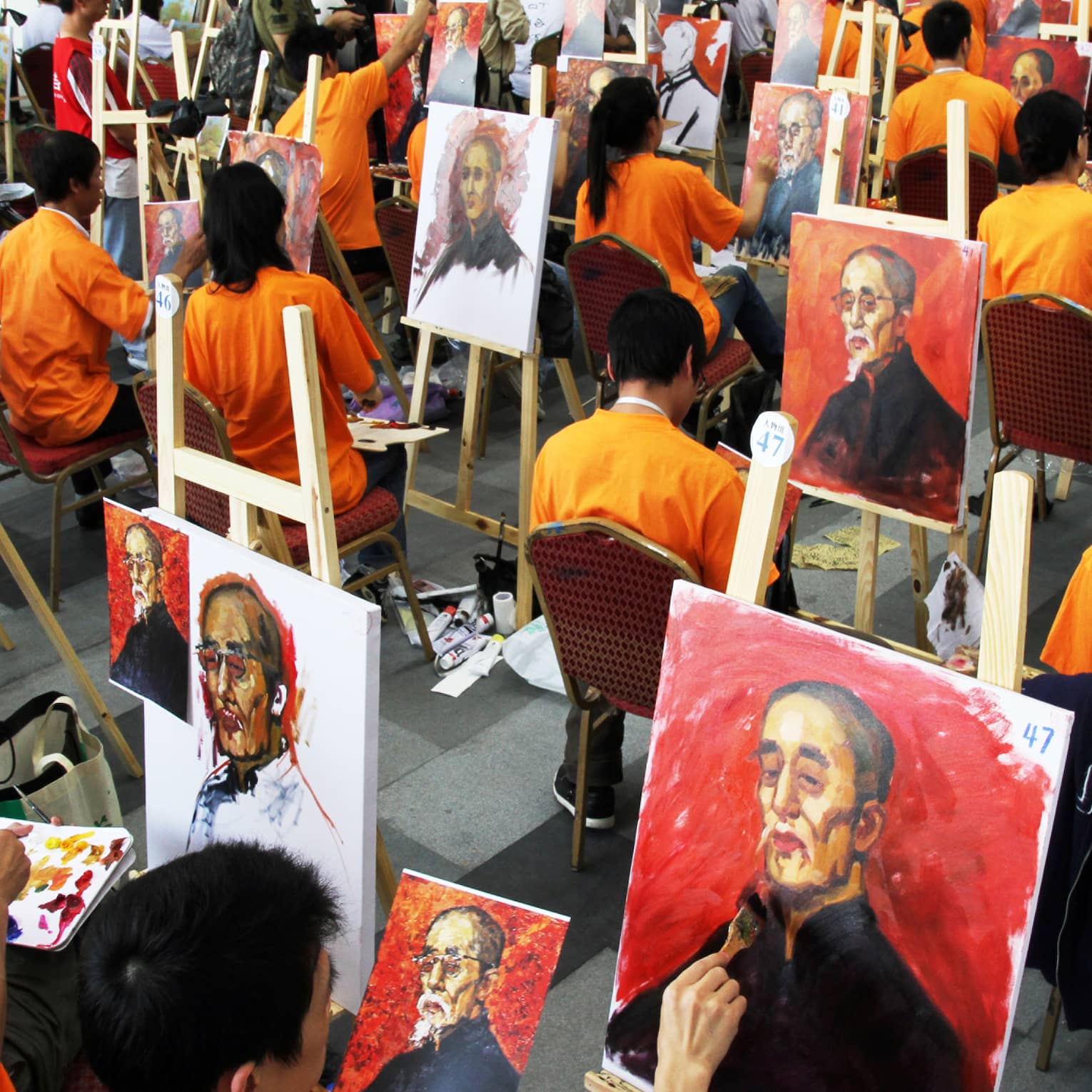 Dozens of people wearing orange shirts sitting at easels and painting the same portrait.