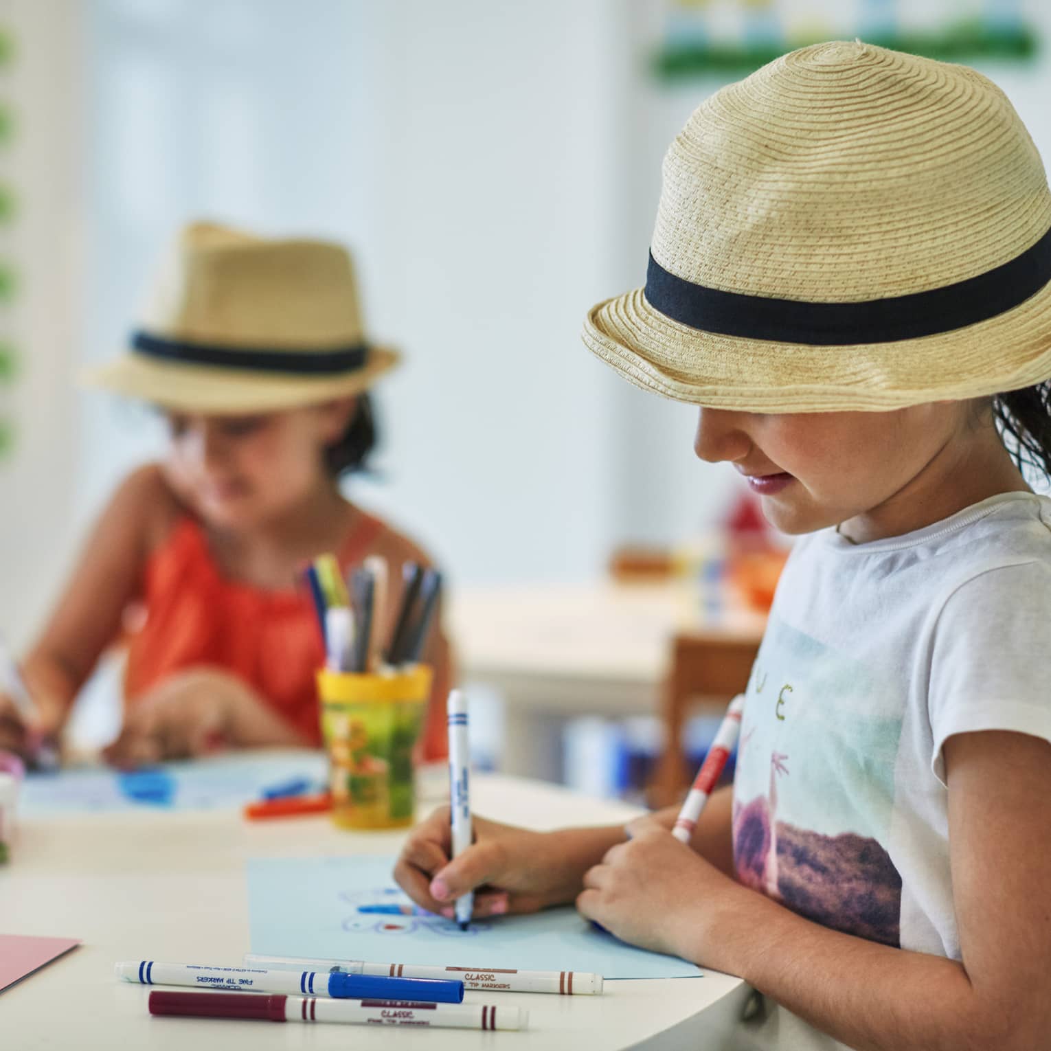 Two smiling children wearing straw fedora hats colour pictures with markers at craft table