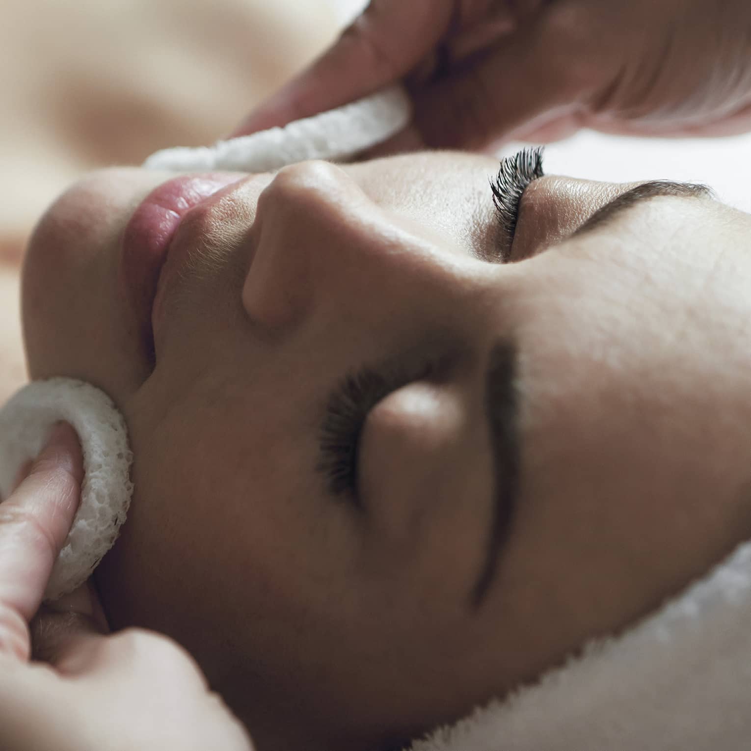 A detail of hands rubbing a sponge on a woman's face as she lies on a massage table with her eyes closed and hair wrapped up in a white towel