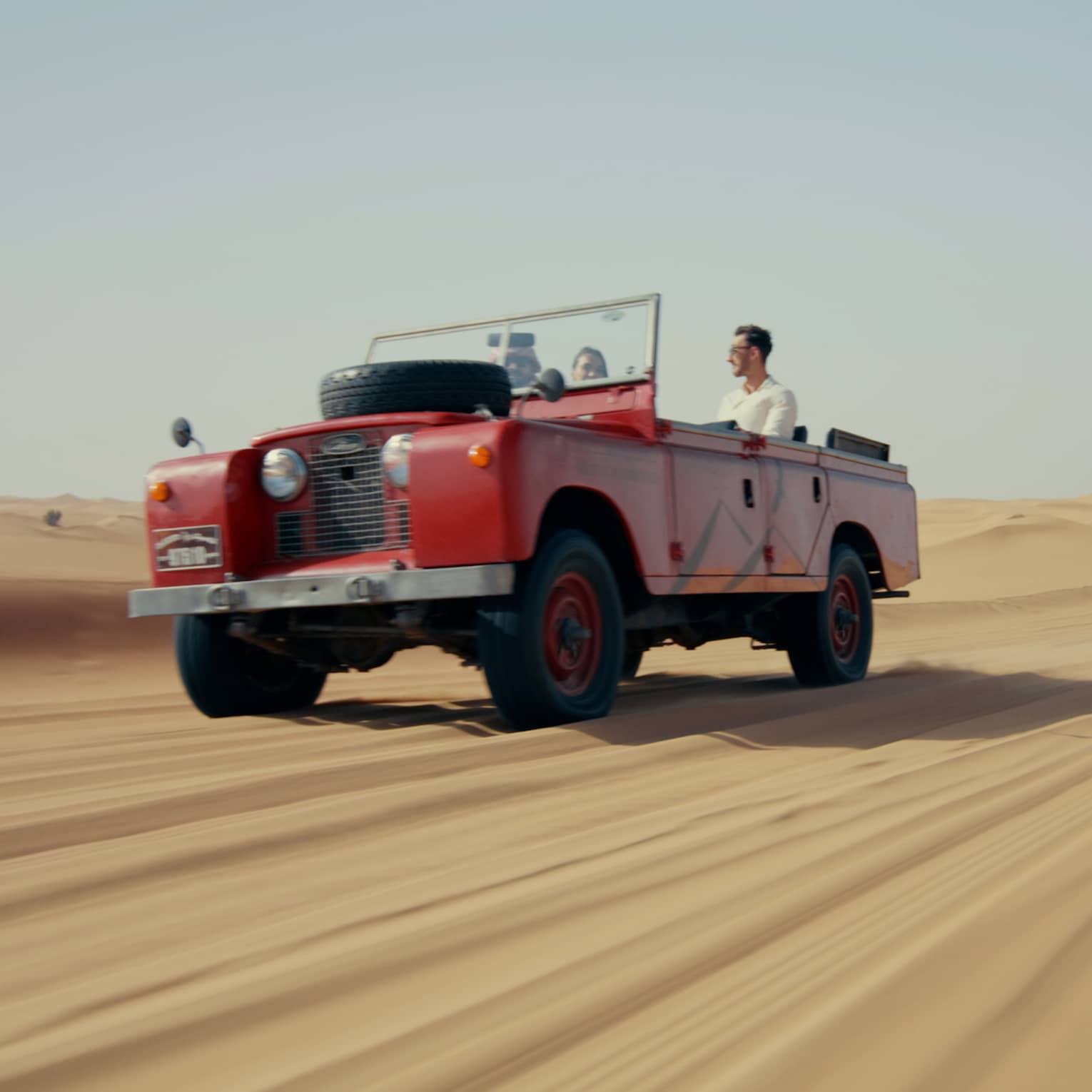 A red truck driving down a long stretch of sand under a blue sky.