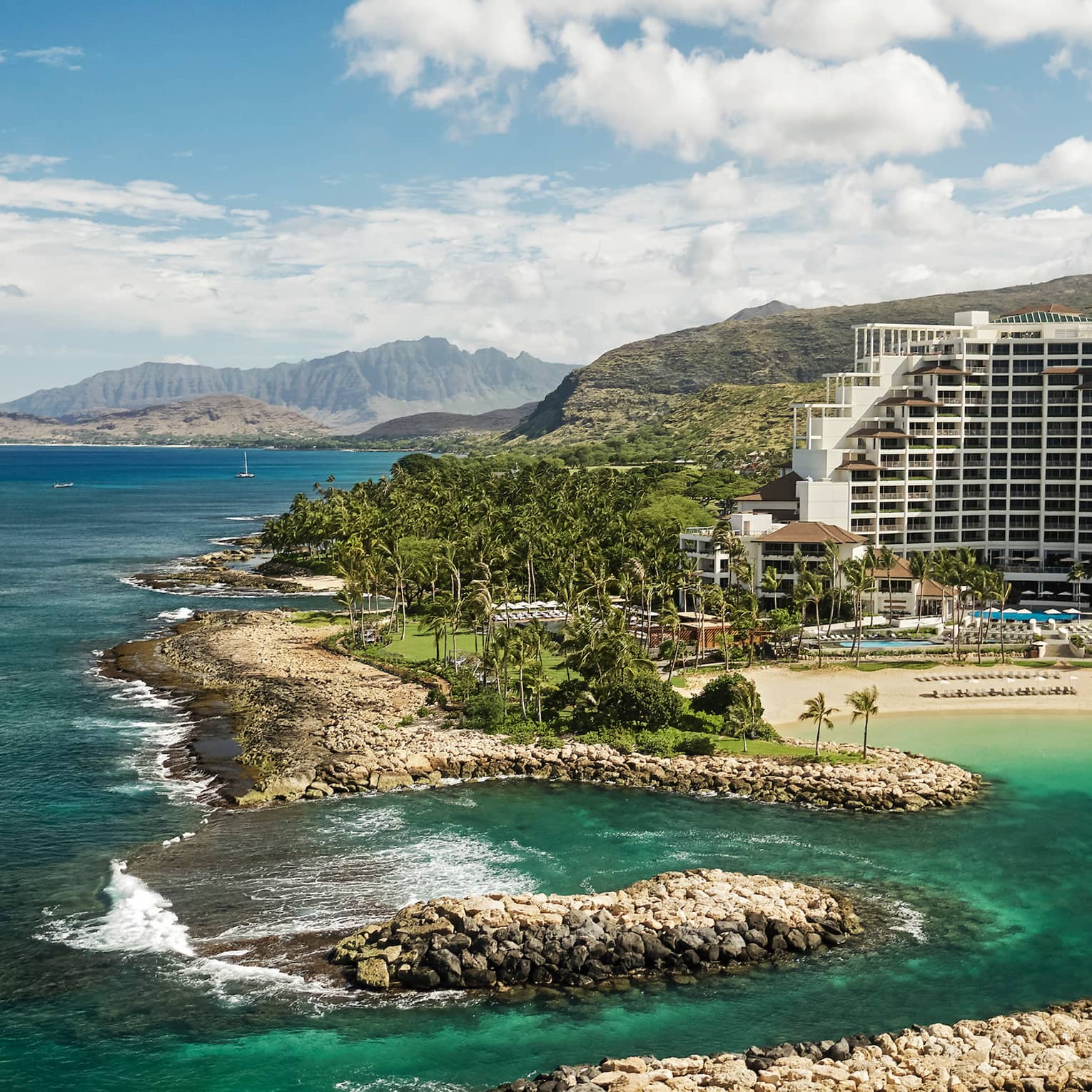 Tall white Four Seasons Resort Oahu building on mountainside by sand beach, rocks, ocean