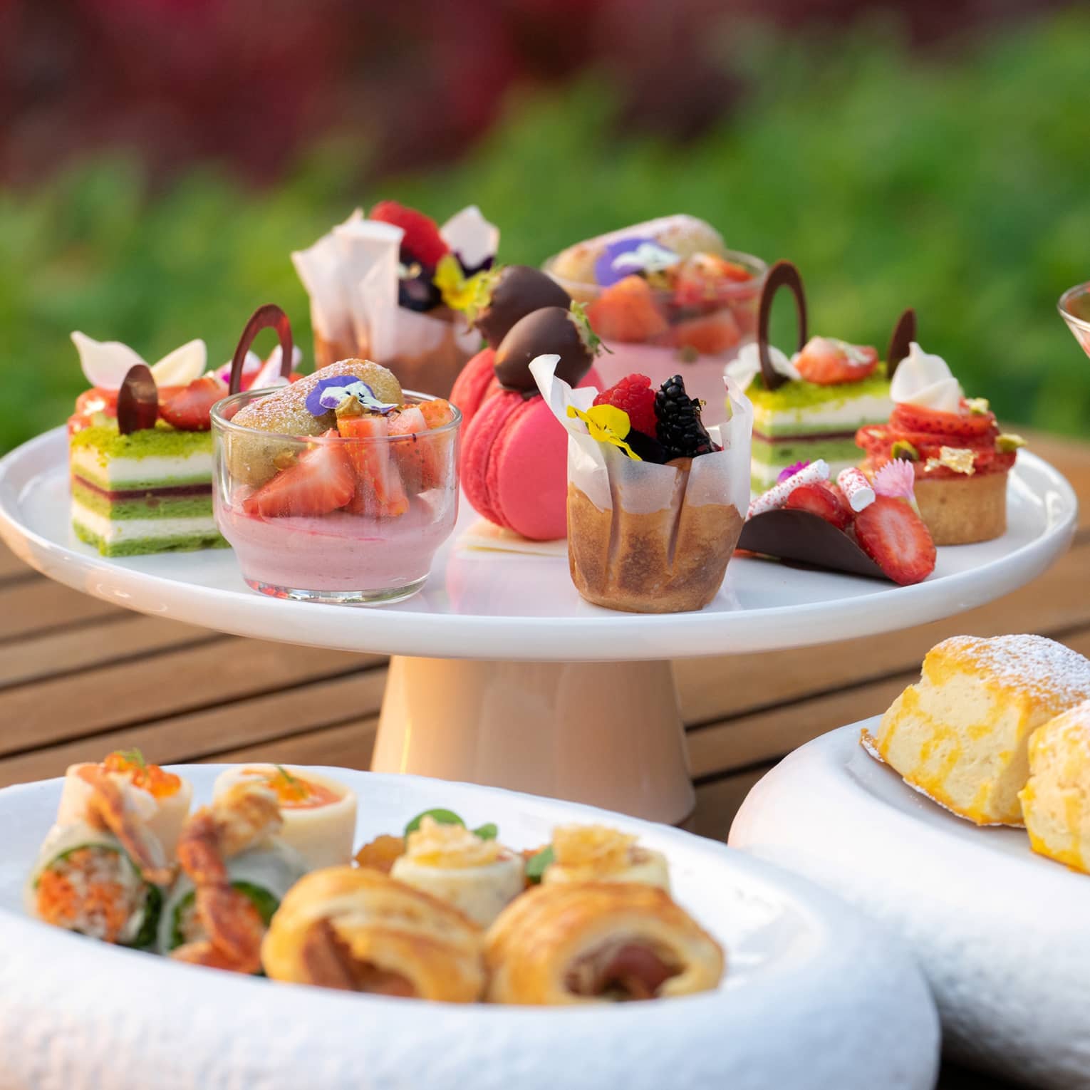 An elegant afternoon tea spread featuring assorted desserts, pastries, scones, and strawberry parfaits on a wooden table in a garden setting.
