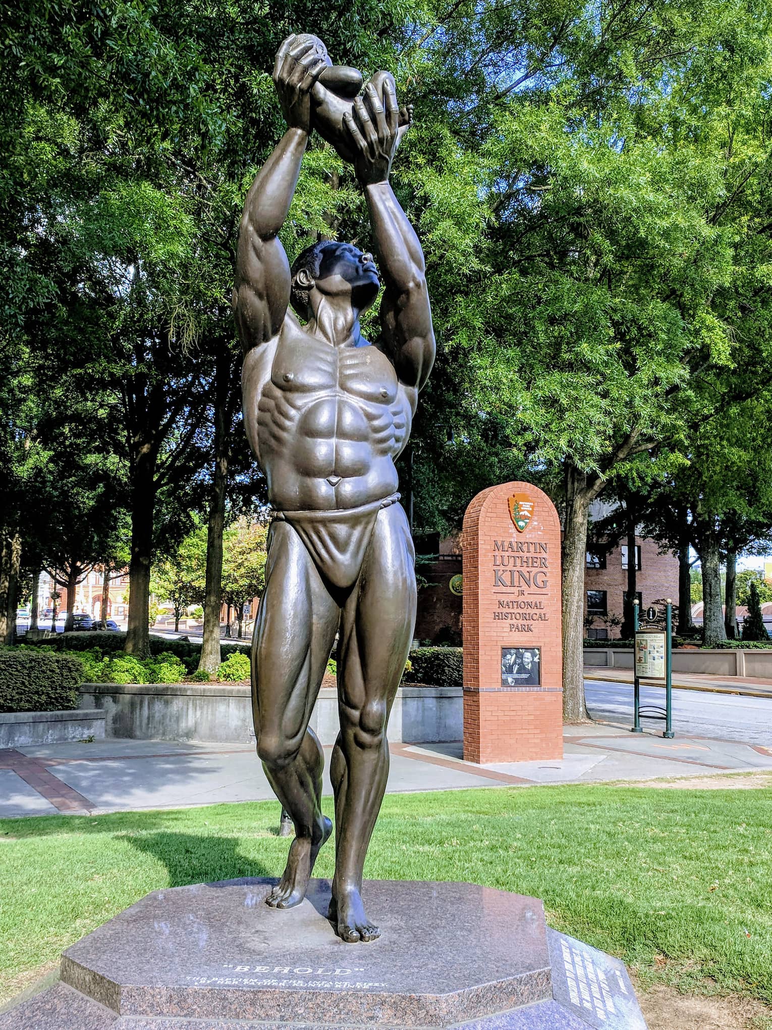 A statue of a man holding something in the air placed in a park with green trees surrounding it.