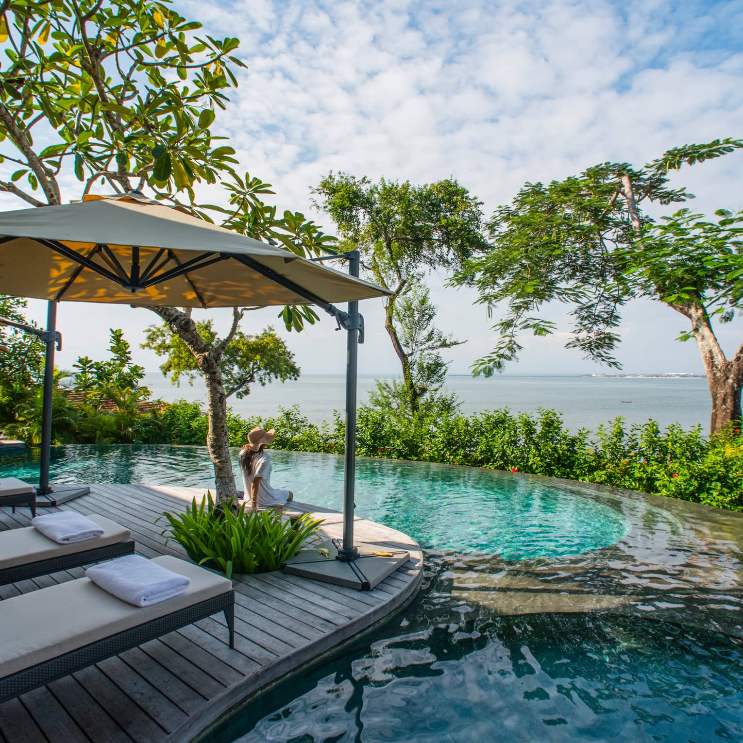 Lounge chairs with umbrella on wooden deck overlooking infinity pool, trees and ocean