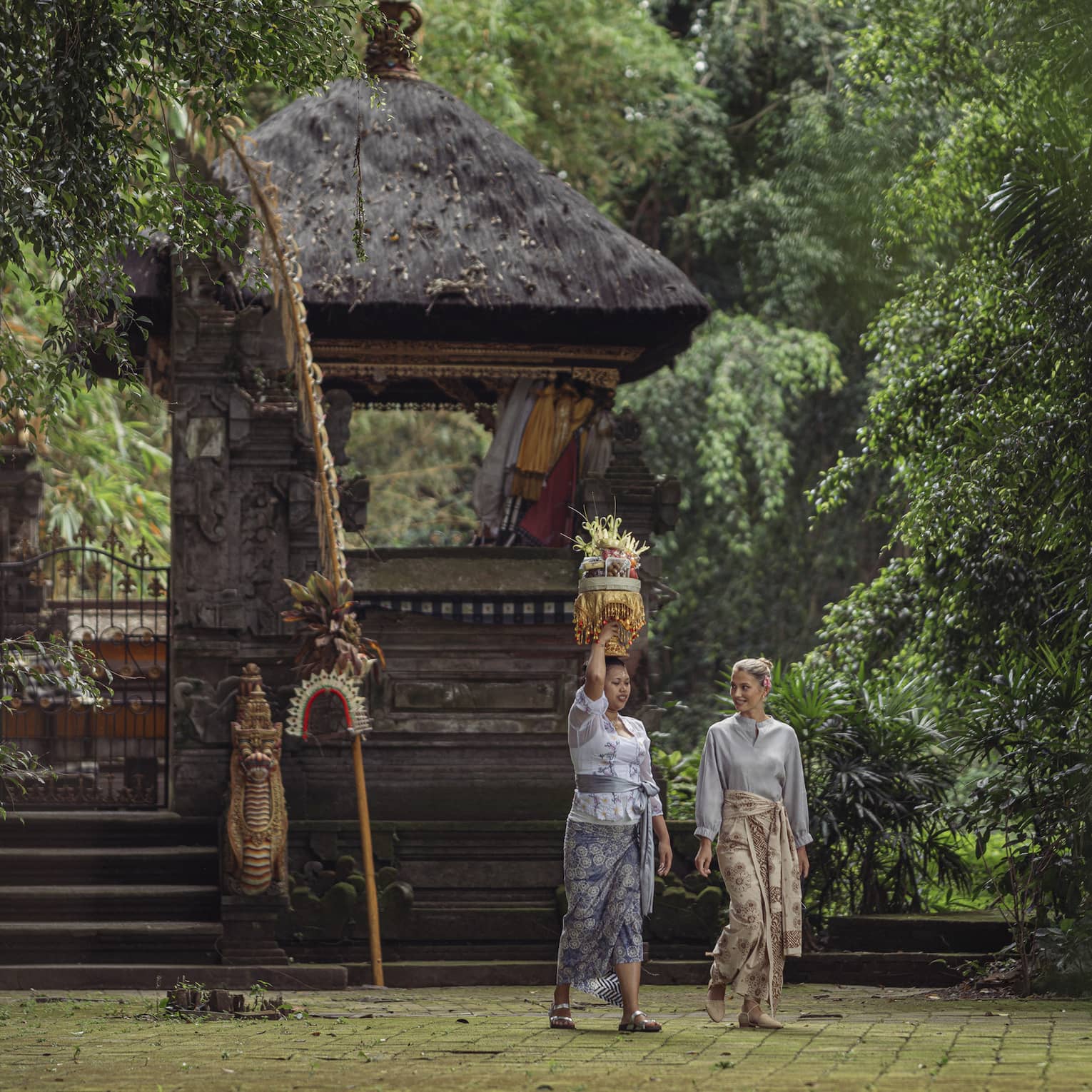 Two women wearing batik skirts walk by a temple in a lush tropical area as one of them carries a golden tray on her head.