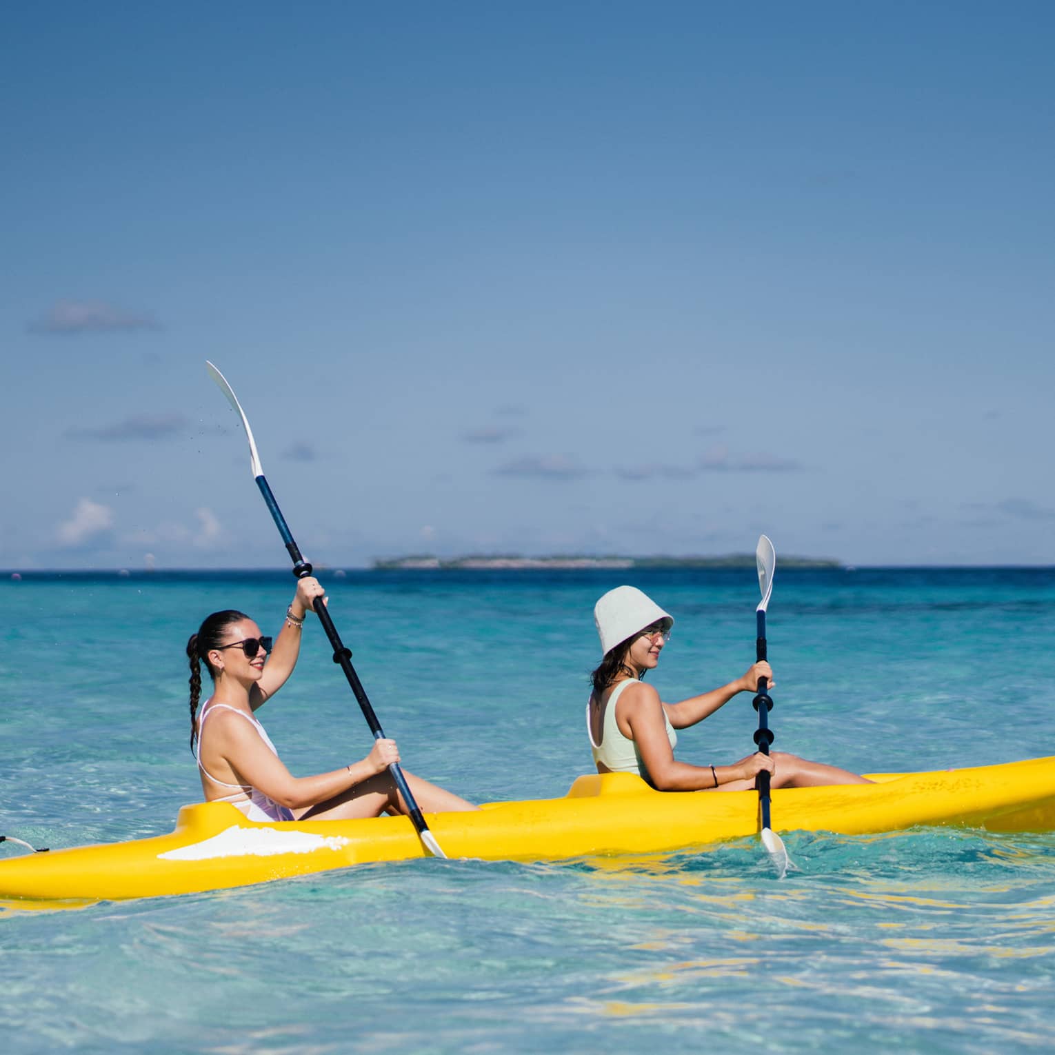 Two people smile as they paddle a yellow kayak in a turquoise lagoon under a clear blue sky; low island in the distance.