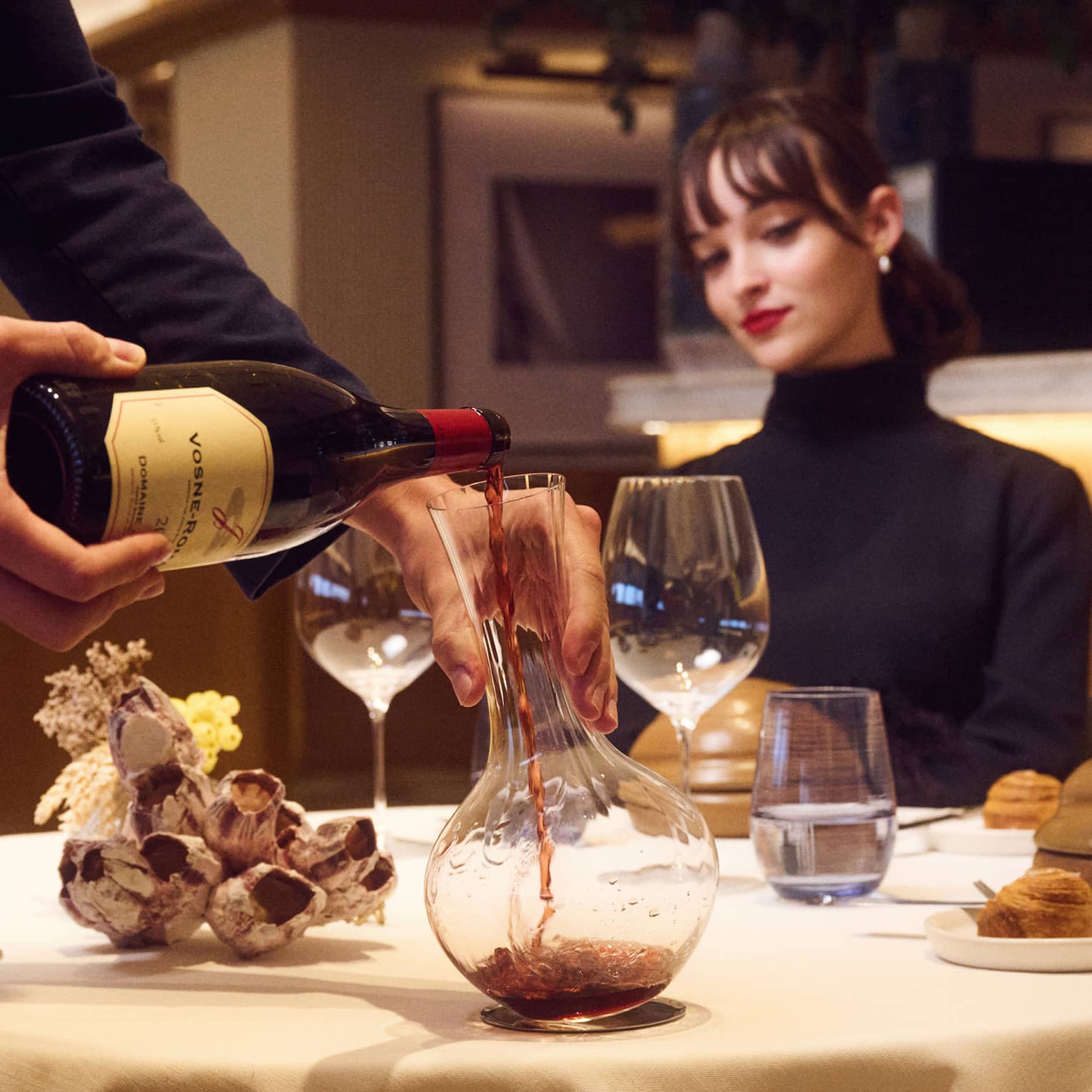 Suited server decants red wine at table with seated guest watching