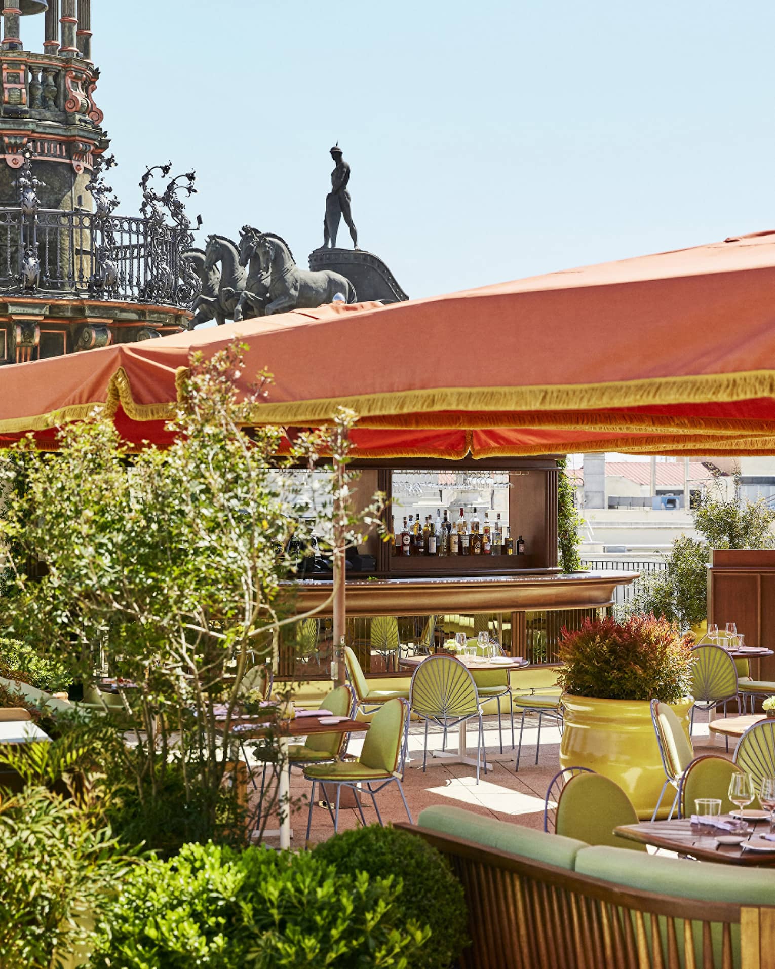 Outdoor terrace brasserie with green chairs and tables under an orange canopy, surrounded by plants, with statues and architectural details in the background