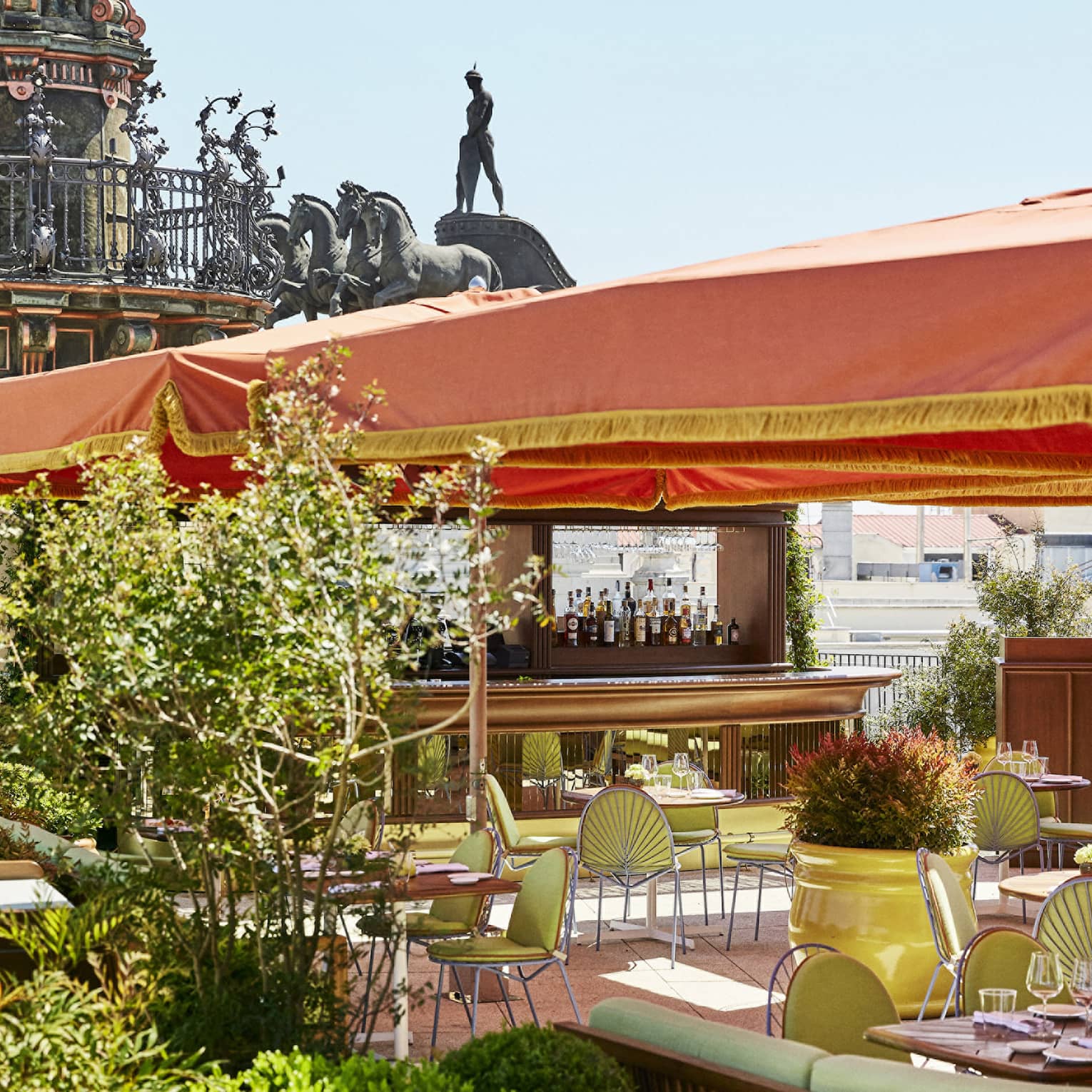 Outdoor terrace brasserie with green chairs and tables under an orange canopy, surrounded by plants, with statues and architectural details in the background
