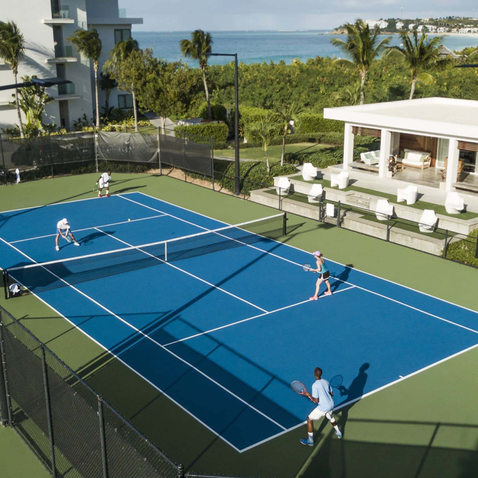 Aerial view of a doubles match on a blue tennis court, with green bordering, palm trees