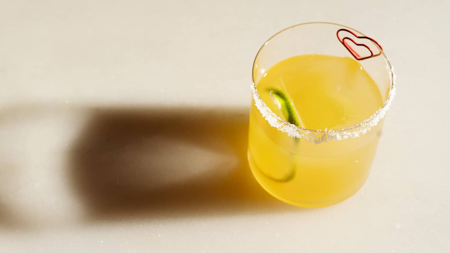 A pale yellow-orange drink in a clear tumbler, with citrus slice garnish and salt rim