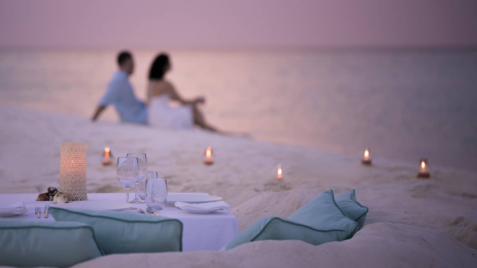 Private dining table in front of sand beach with tealight candles, couple sitting in sand