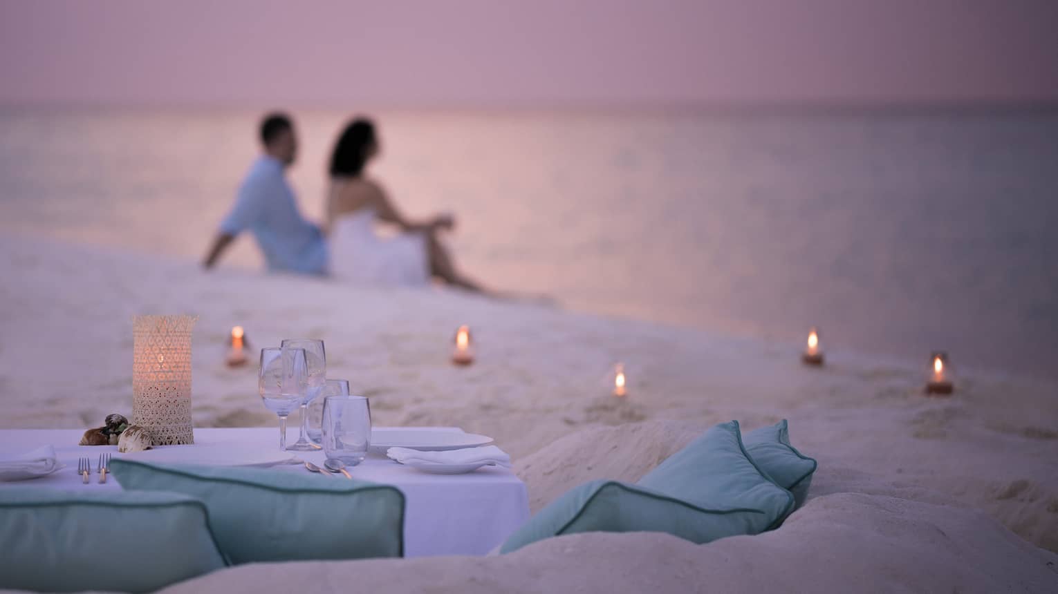 Private dining table in front of sand beach with tealight candles, couple sitting in sand
