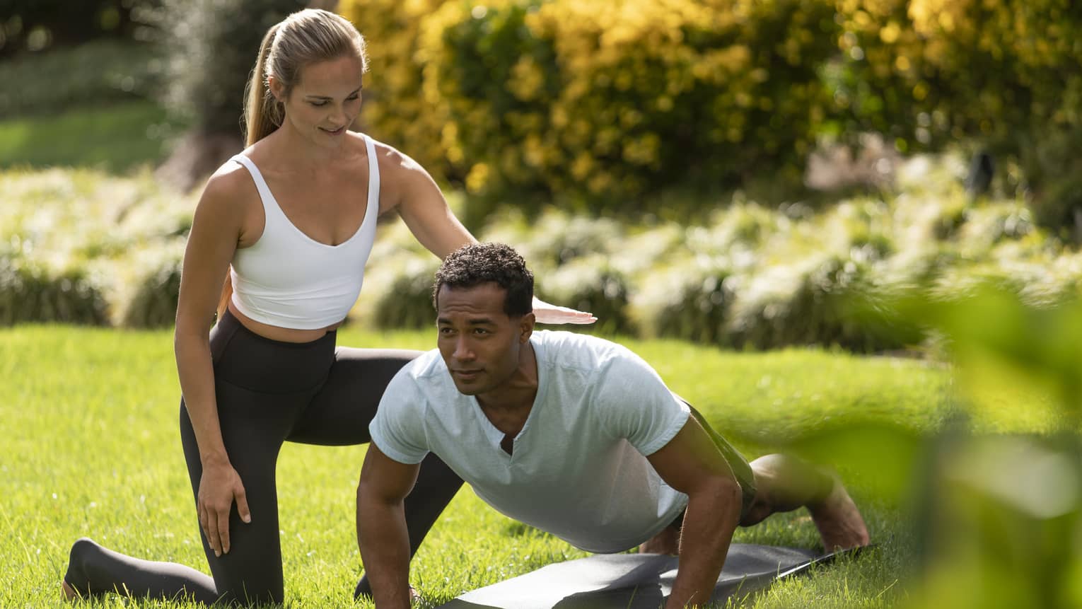 A yoga instructor helping a man get into a yoga position outside on very green grass.