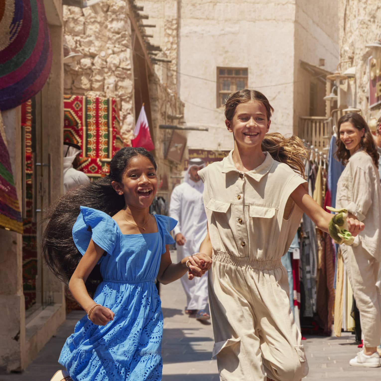 A busy, sunny open-air market between sandstone buidlings; two children run among shoppers browsing vibrant clothing.