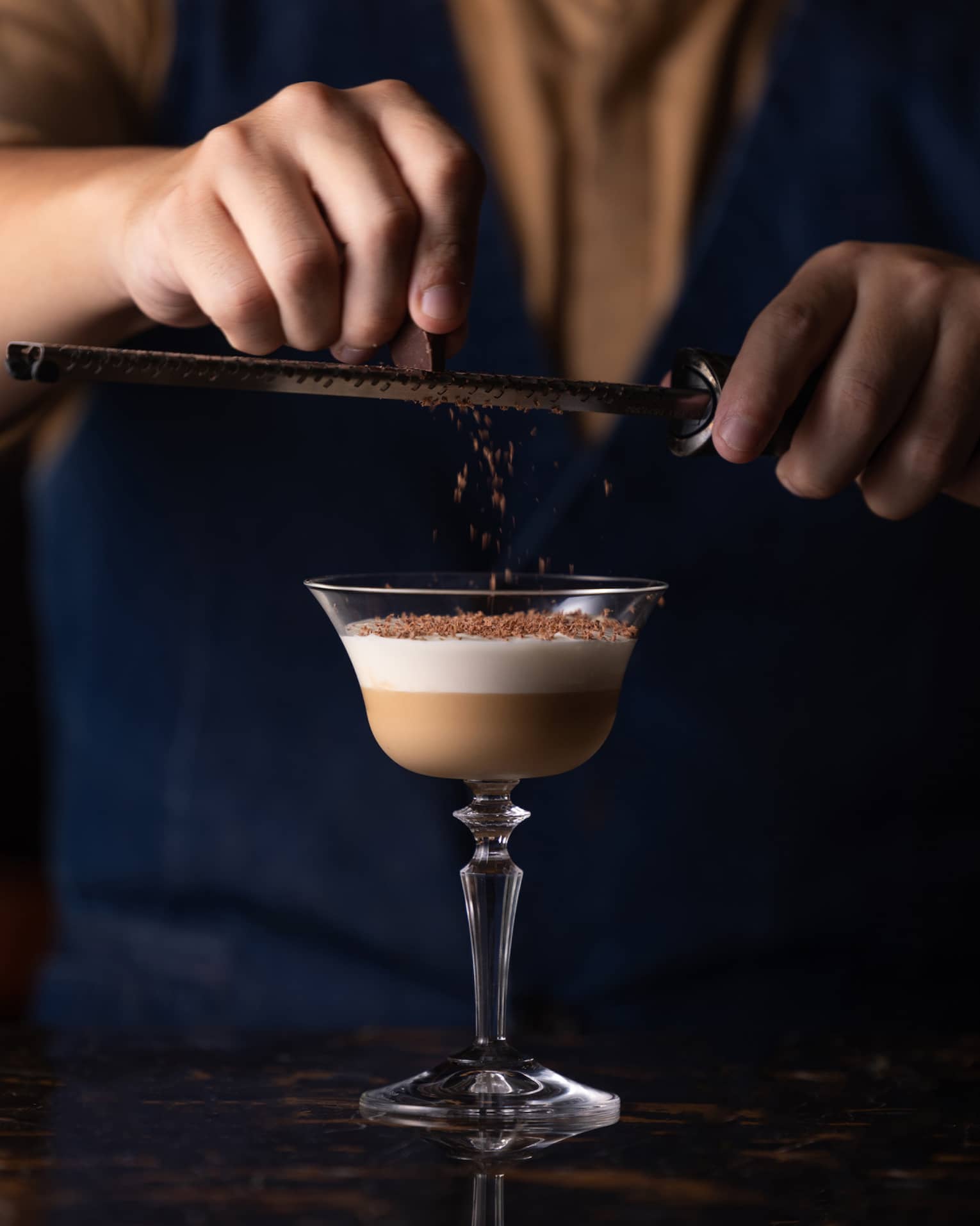 Close-up view of person holding a grater above a cocktail glass and garnishing its two-tiered contents with chocolate shavings