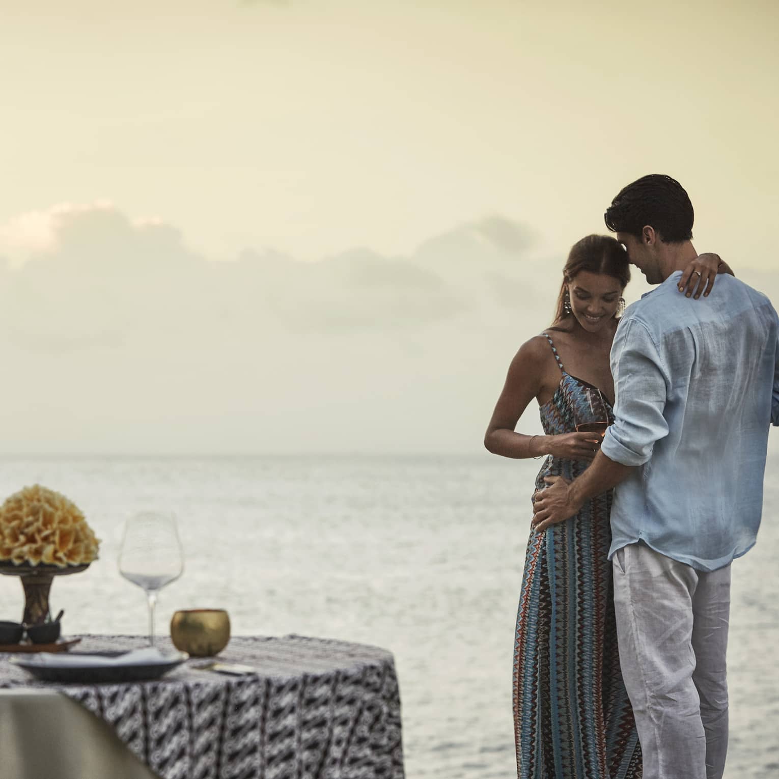 Couple embraces beside private dining table overlooking ocean at sunset