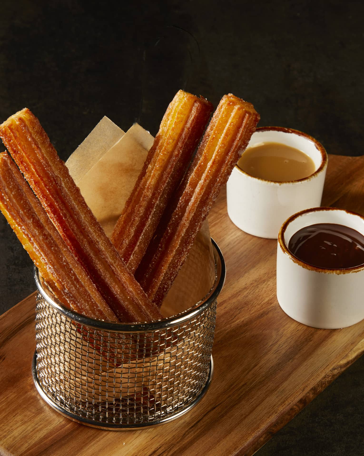 Crispy, golden churros coated in sugar in a metal basket on a wood board with chocolate and dulce de leche dipping sauces. 