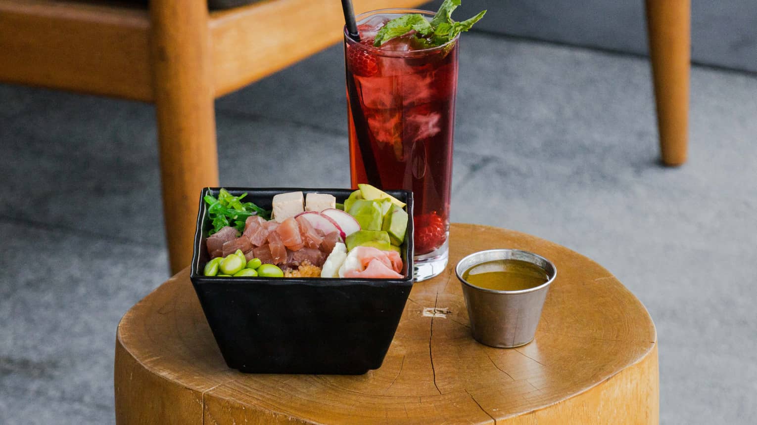 A black square bowl filled with poke, greens and an assortment of vegetables sits next to a red-colored beverage in a tall glass, both on a small round wooden table