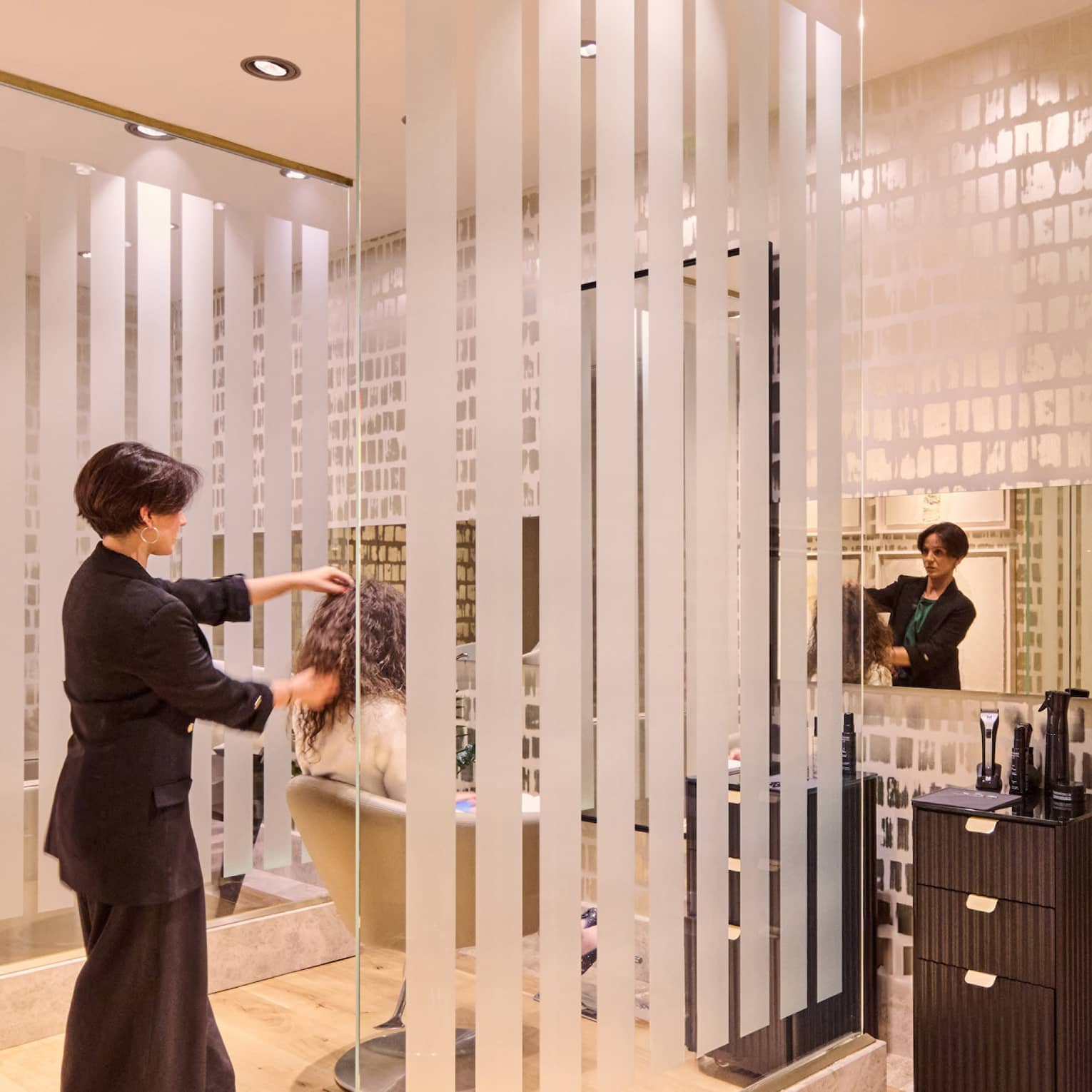 A hair stylist styles a guest?s hair in a brightly lit salon with pale wood floors, large mirrors, salon chairs and contemporary decor.