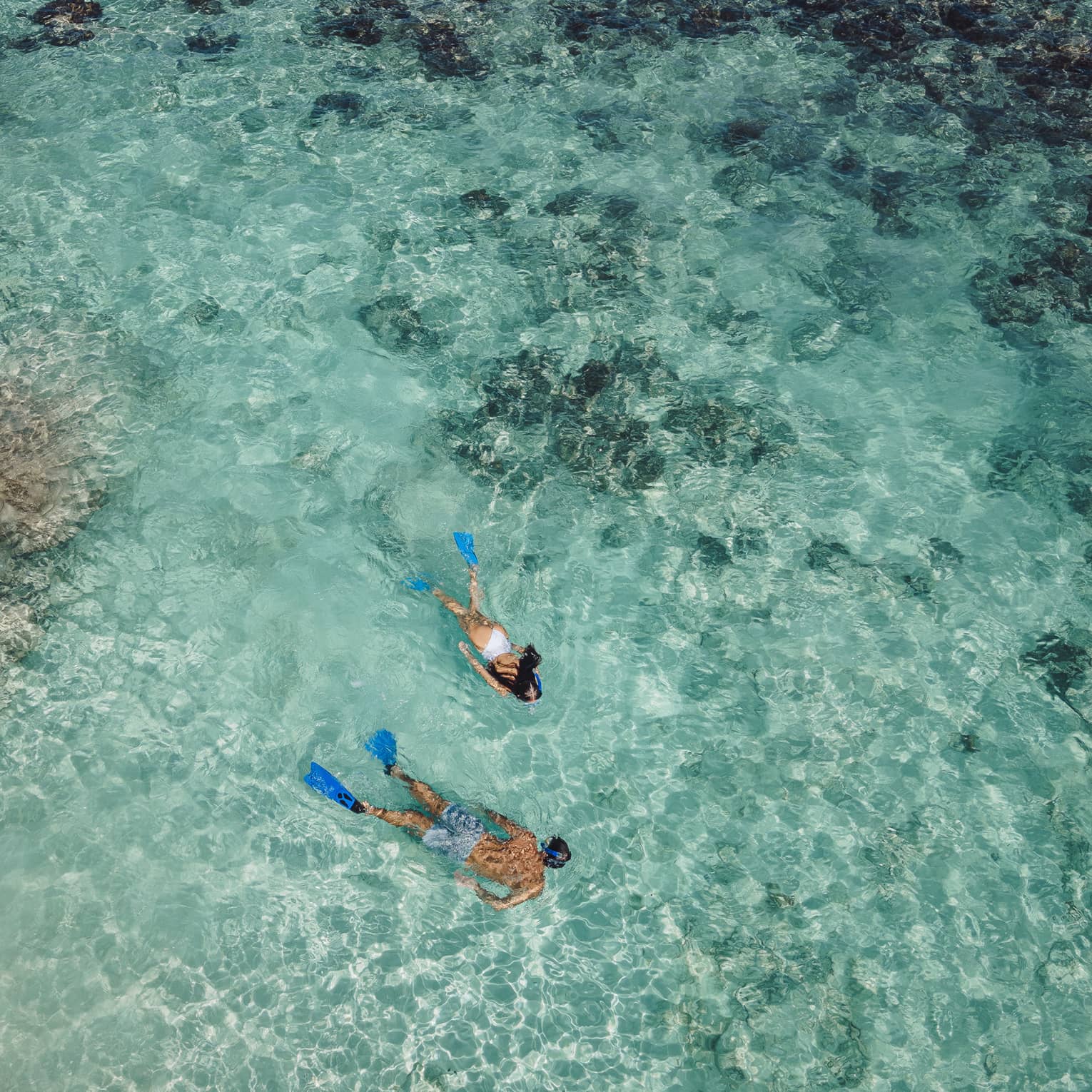 Aerial view of two snorkellers wearing blue flippers in an expanse of shallow, turquoise waters rippling over the reef.