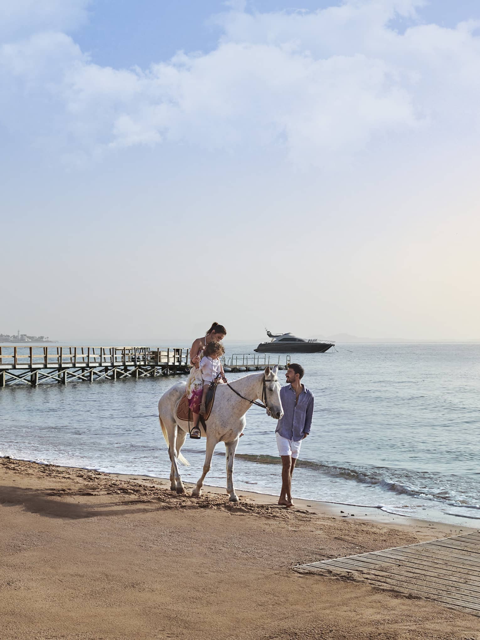 An adult and child on a white horse on a beach, led by another guest, with a wooden pier and a yacht visible in the distance.