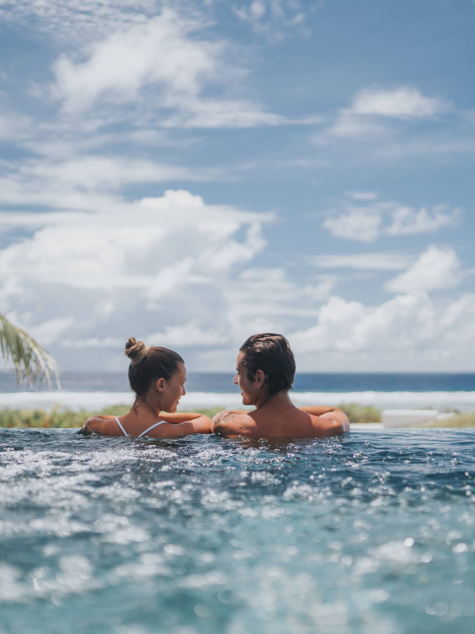 Back of couple at edge of whirlpool overlooking ocean, blue sky 