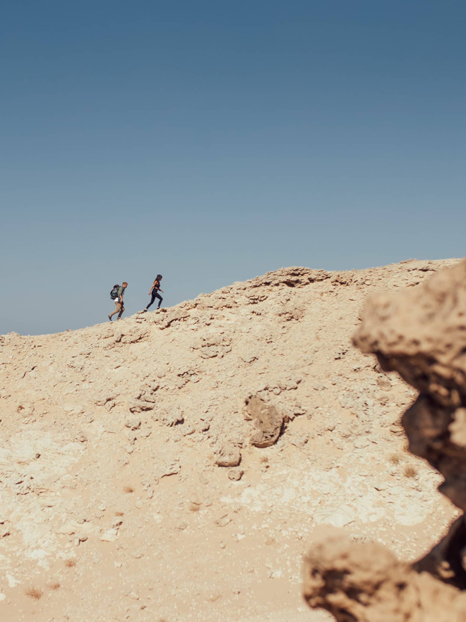 Against a background of sunny, clear blue sky and surrounded by rocky terrain, two people hike up a sandy hillside.