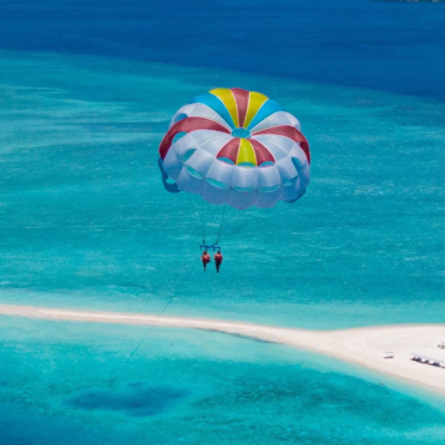 Aerial view of two people parasailing in tandem, colourful sail floating above an expanse of aqua water and a white sandbar.