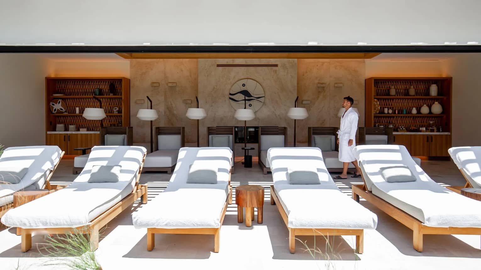 A man dressed in a white spa robe walks in an outdoor spa pavillion behind a row of white lounge chairs
