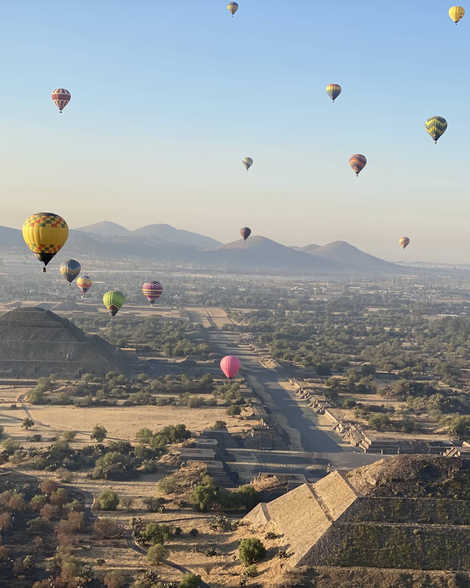 An aerial view of colourful hot-air balloons floating over pyramids and trees, with mountains and a city in the background.