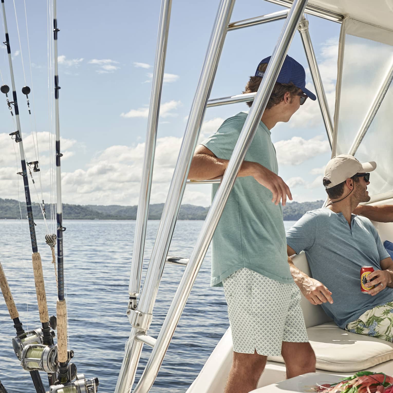 Aboard a fishing vessel on calm water, three people relax under the shaded cockpit, their fishing rods stored beside them. 